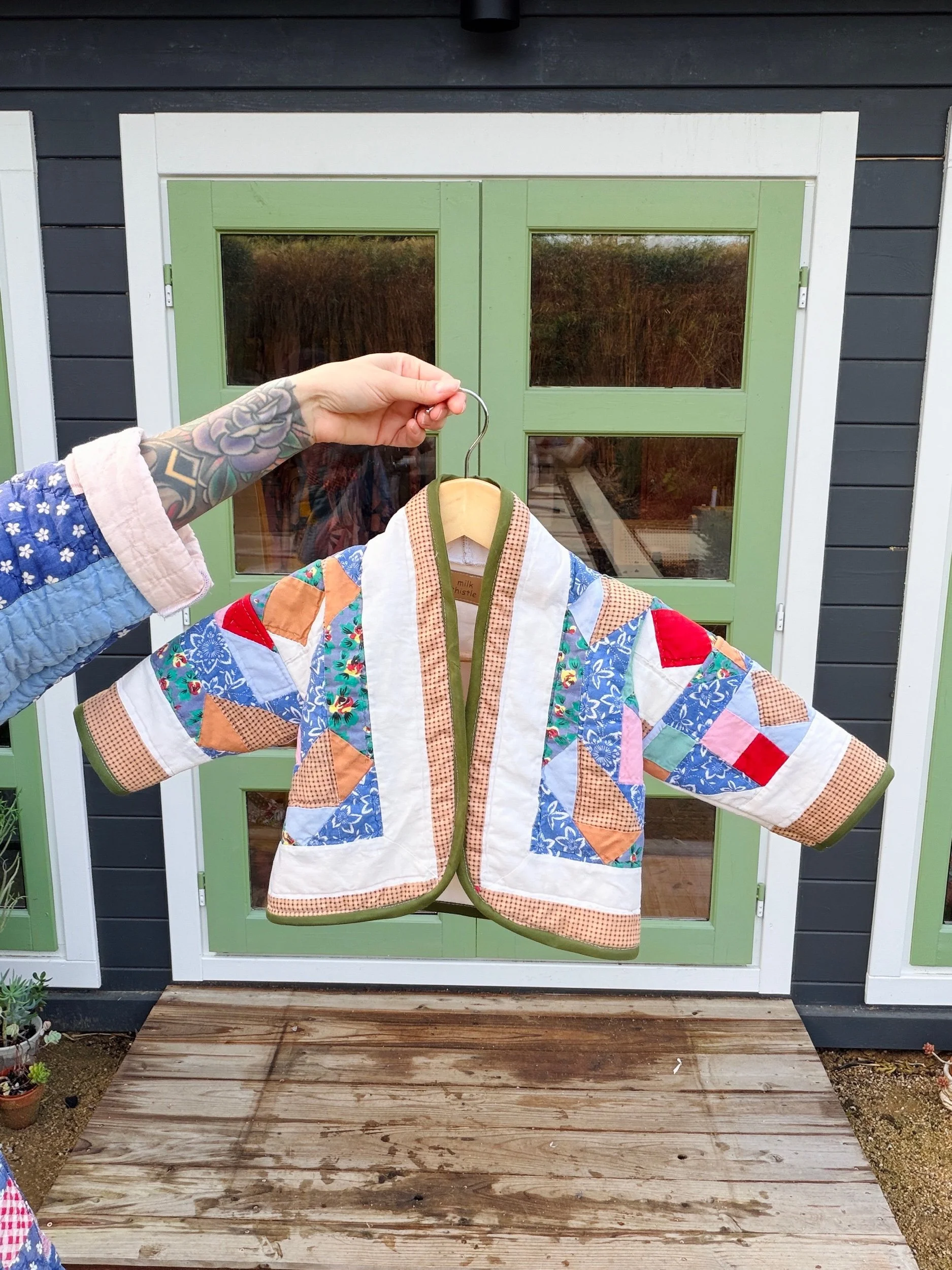 Person holding a colorful patchwork quilted jacket on a hanger in front of a green and white windowed door.