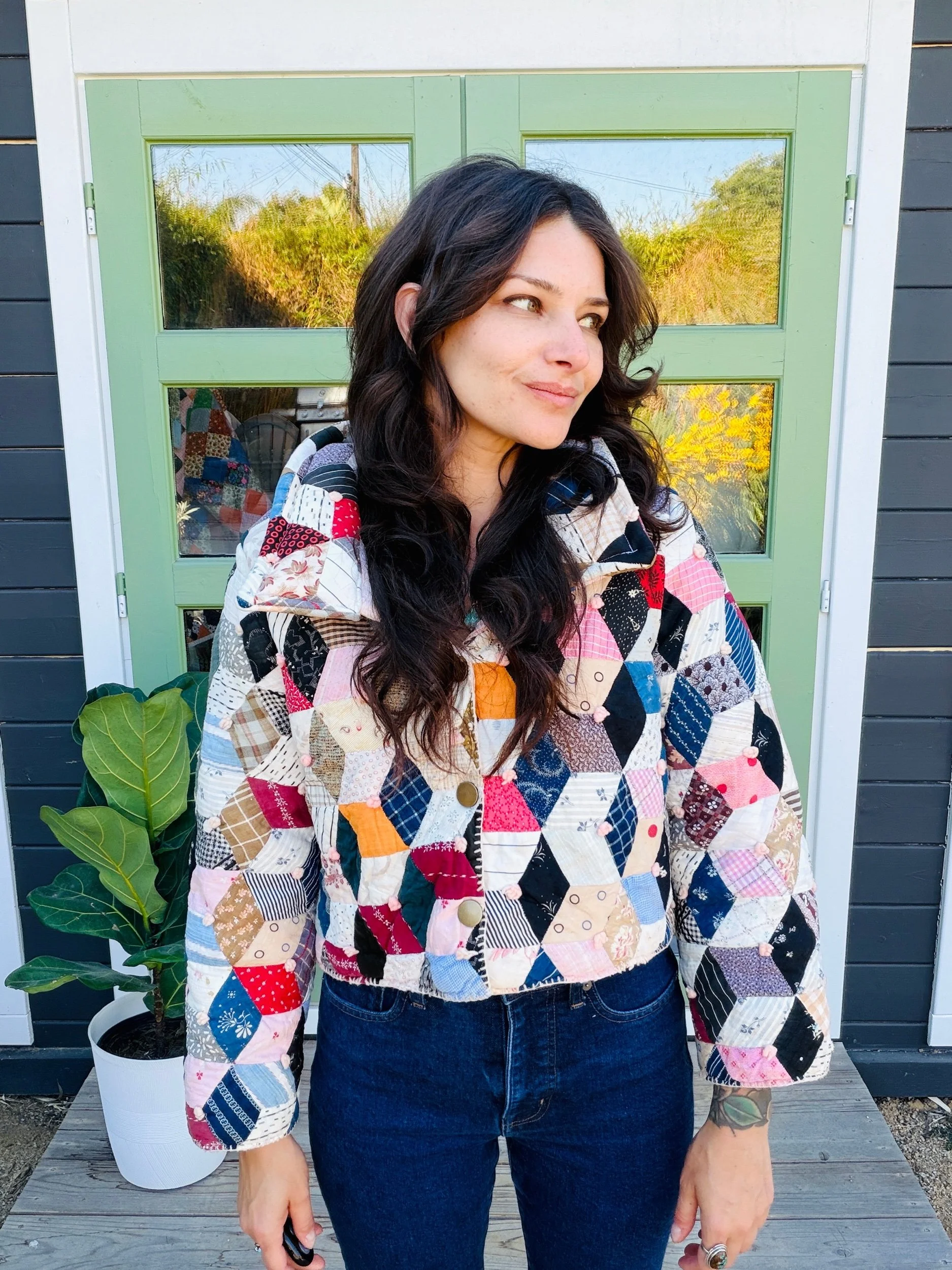 A woman with dark wavy hair wearing a colorful patchwork jacket and dark jeans standing in front of a green door with reflective glass panels, next to a potted plant with large green leaves.