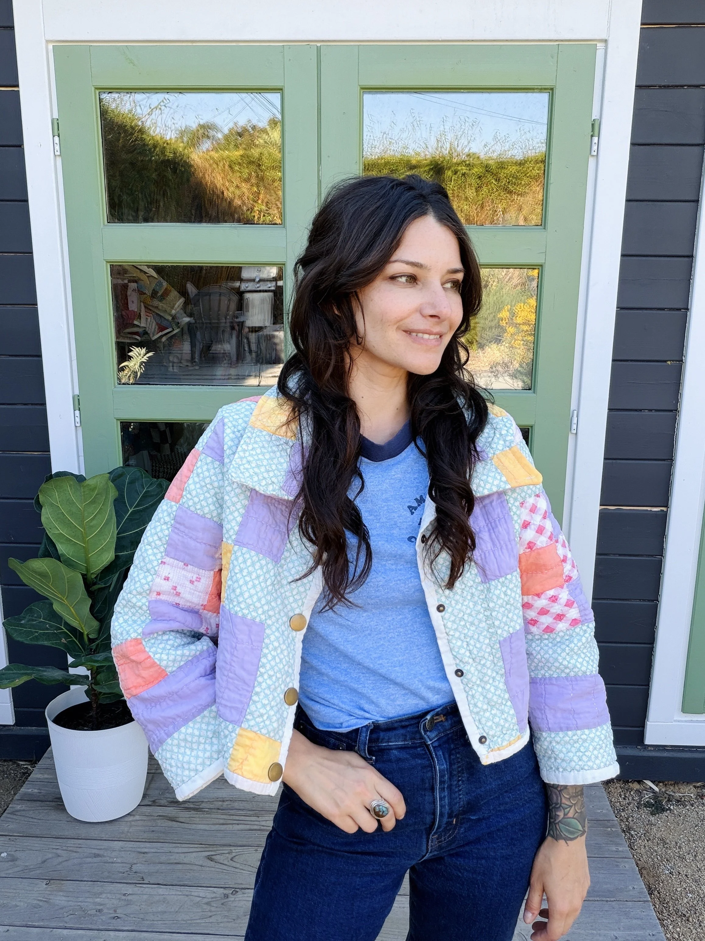 A woman with dark hair and tattoos on her left arm standing outside in front of a green door with windows, smiling with her head turned slightly to the left, wearing a patchwork jacket over a gray t-shirt and dark jeans.