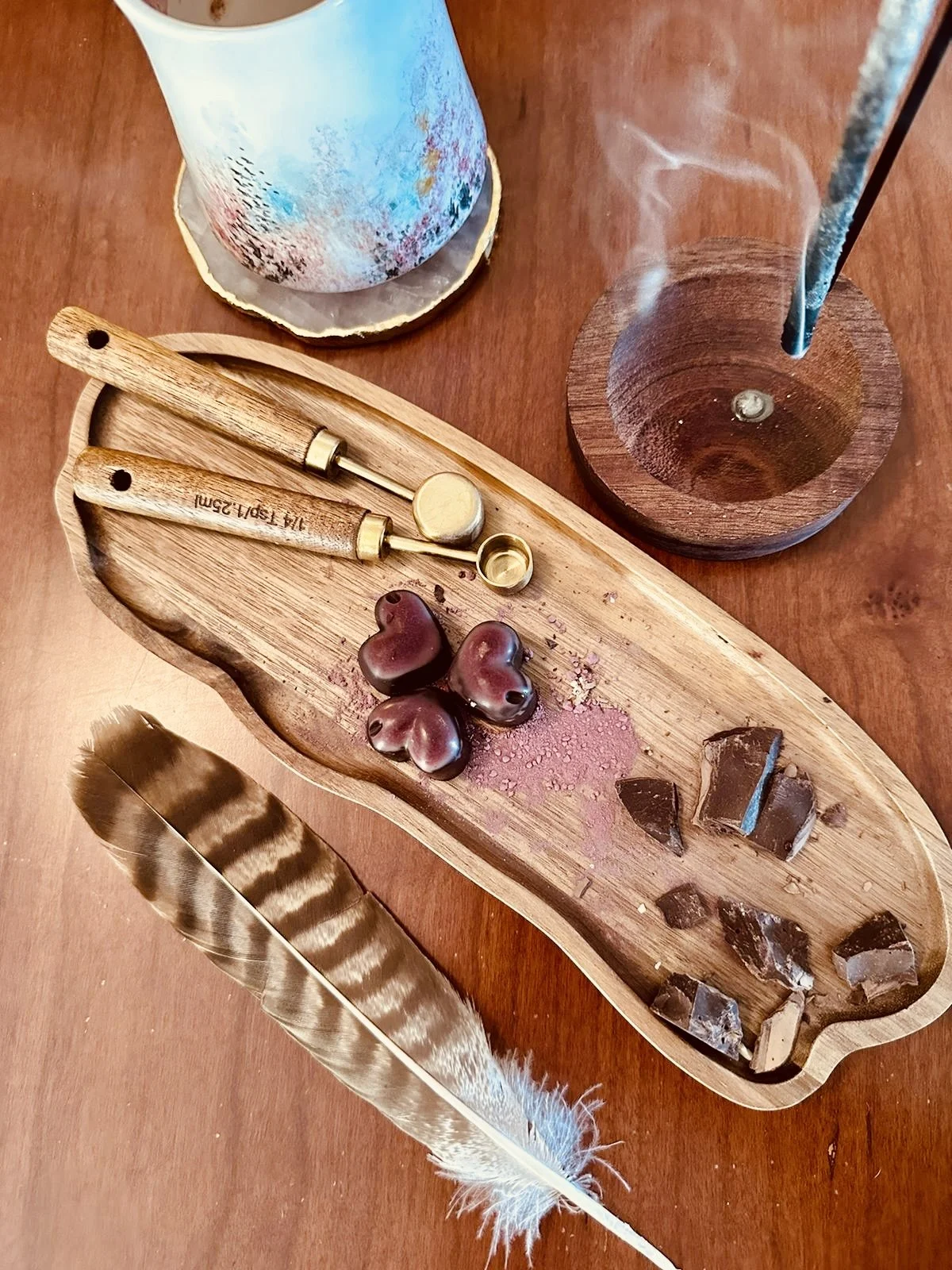 A wooden tray with various stones and two small glass bottles, a large feather, and a lit stick in a holder, with a cylindrical object in the background on a wooden surface.