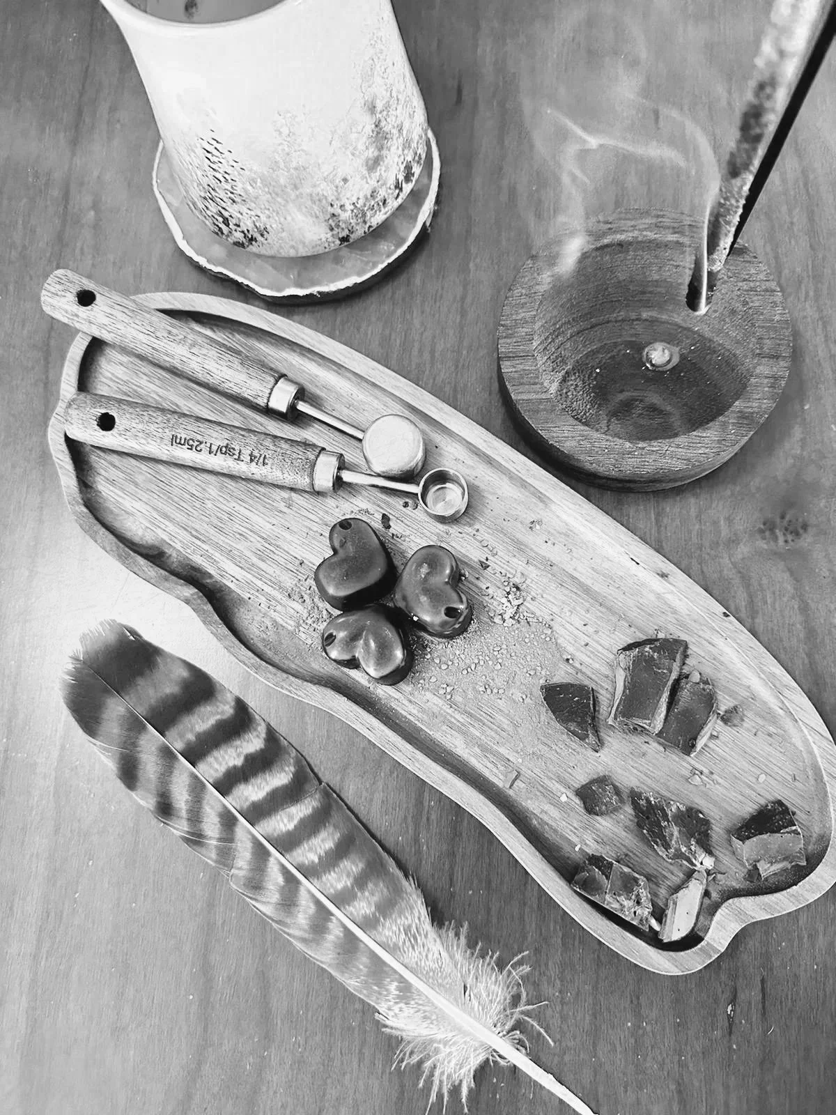 A wooden tray with various stones and two small glass bottles, a large feather, and a lit stick in a holder, with a cylindrical object in the background on a wooden surface.