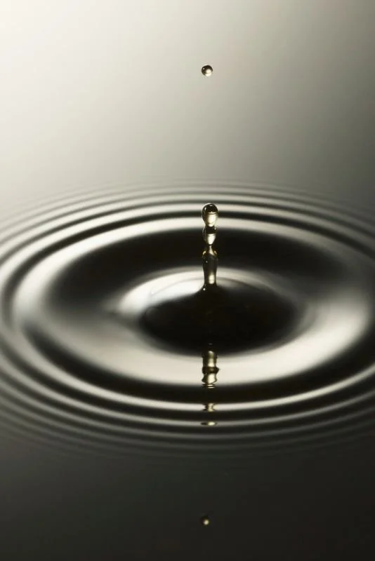 A close-up of a water droplet creating ripples on a smooth water surface with a small droplet in the background.