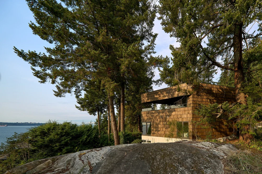 Shingle-clad Rose house among trees and exposed rock, with water beyond.