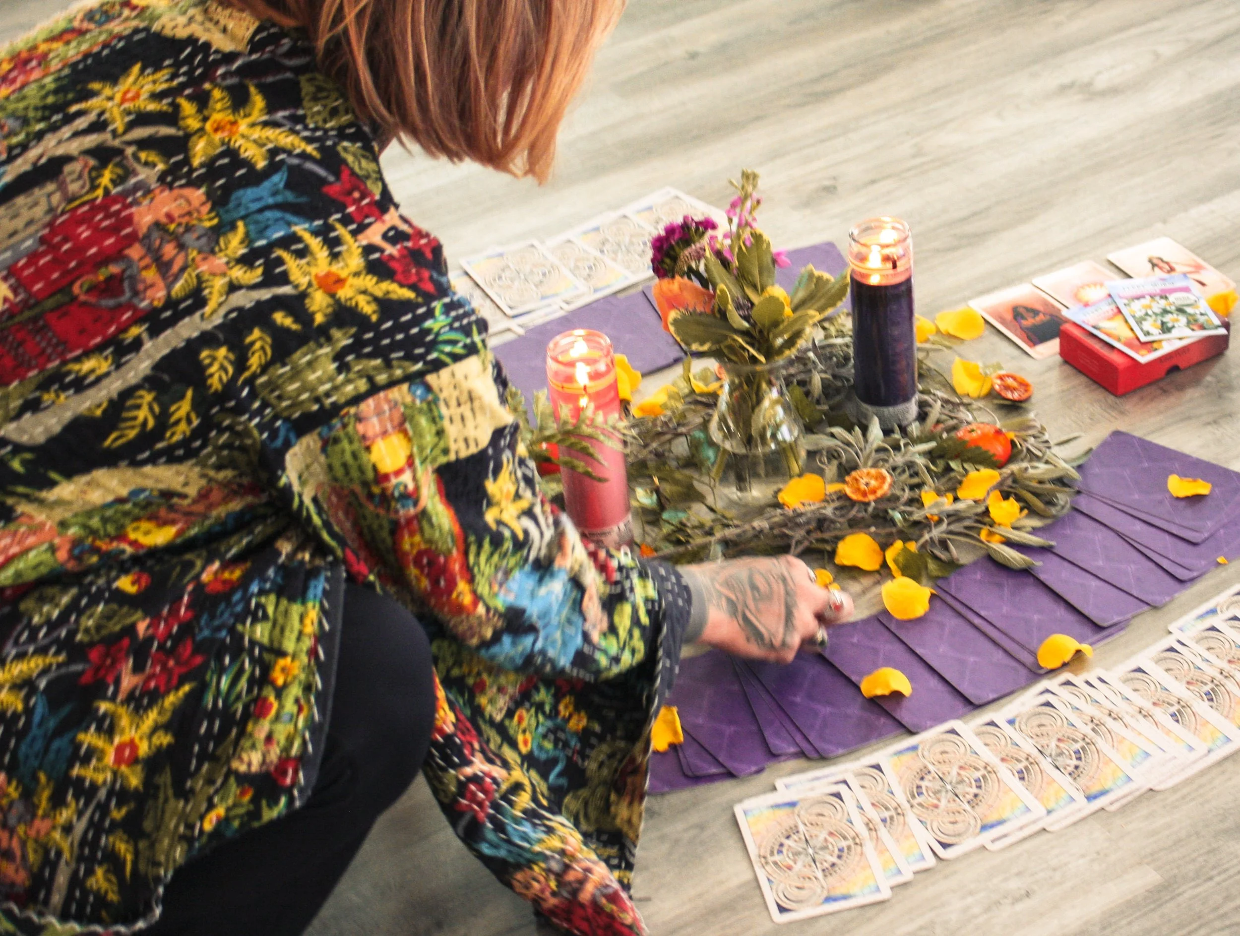 A person with tattoos wearing a colorful embroidered jacket arranging tarot cards on the floor, with candles, flowers, and scattered yellow petals.