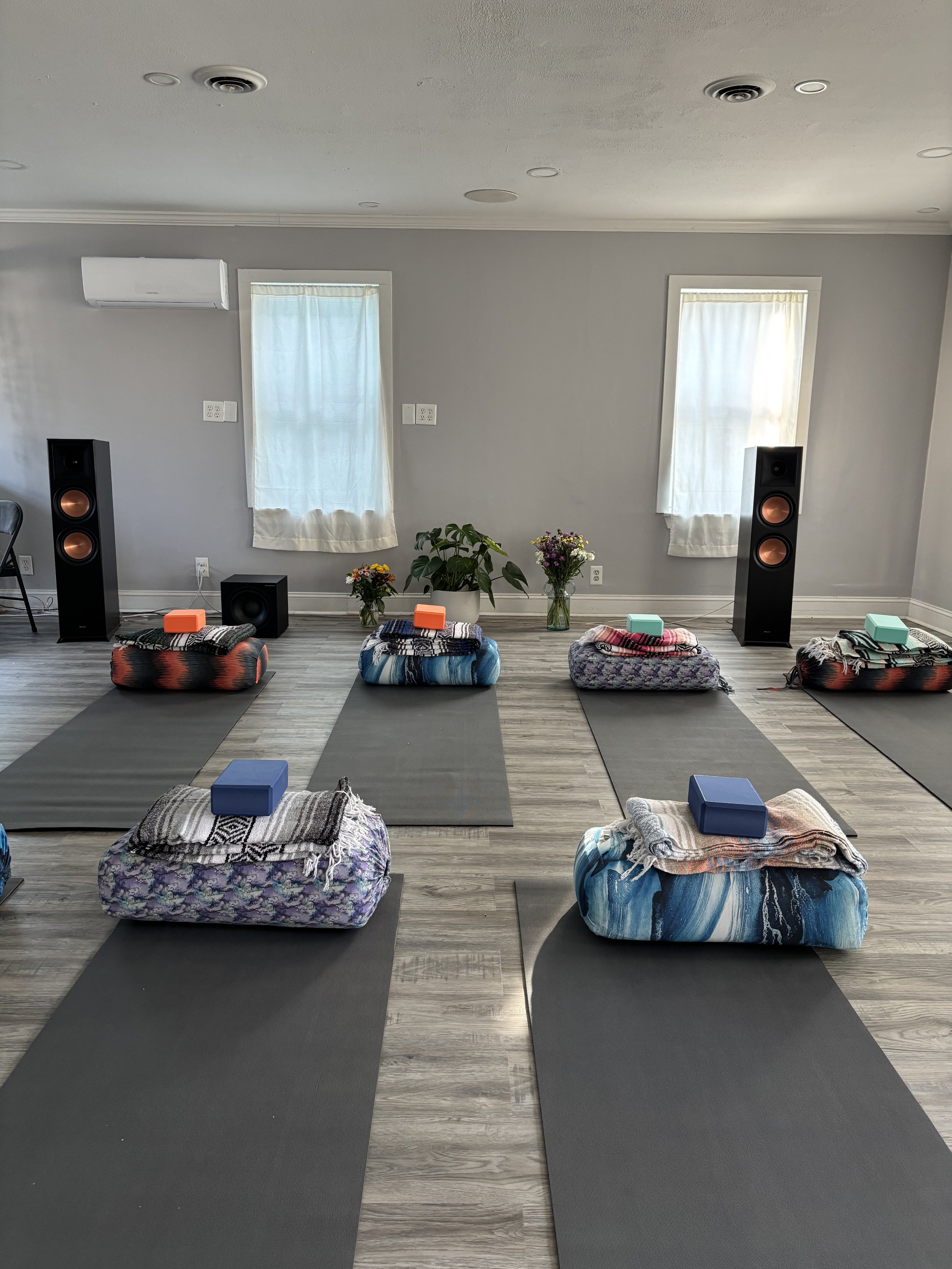 Yoga mats and blankets arranged in a classroom setting with large speakers, potted plants, and small colorful blocks on each blanket.
