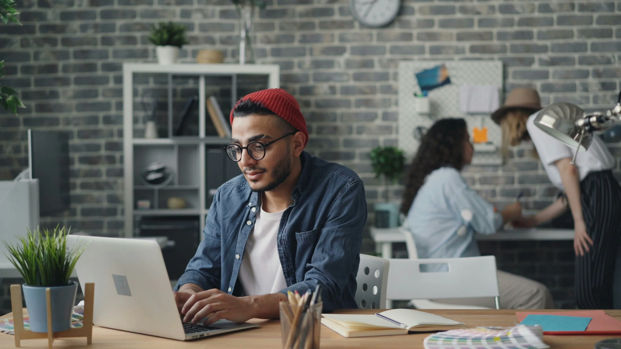 A creative VA wearing a red beanie and denim shirt working on a laptop at a desk with colored pencils, notebook, and plant, in a stylish shared workspace with shelves and a bulletin board