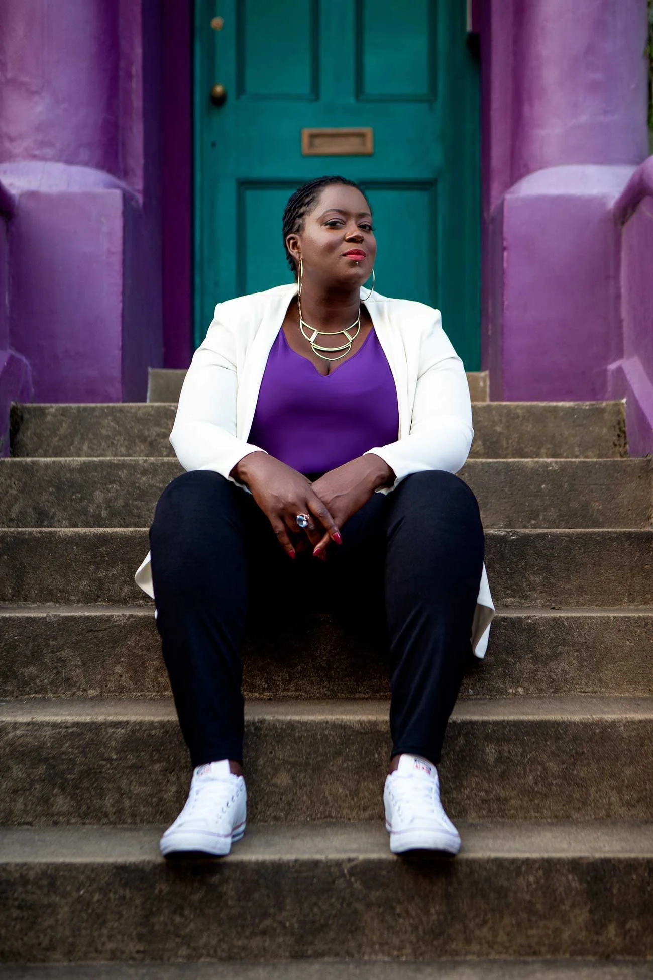 Beautiful black woman chin confidently tilted upwards, sat on the stoop belonging to a bright teal door and purple wall.
