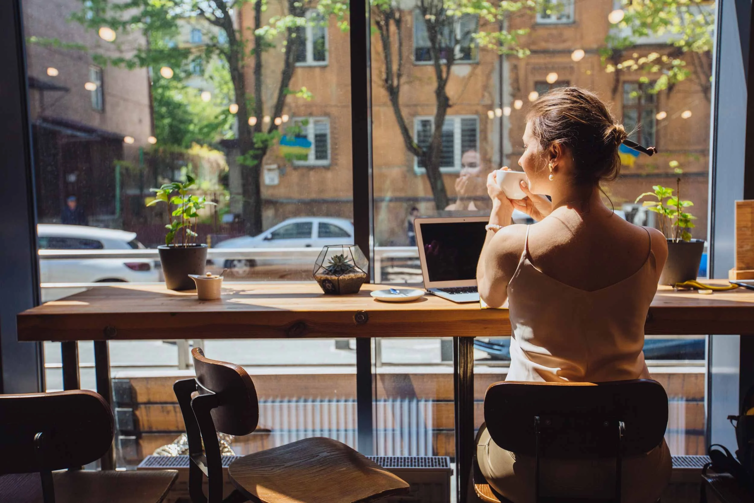 A VA at a café counter facing a large window, holding a cup and looking outside, with a laptop, smartphone, and plant on the counter in a bright, peaceful workspace