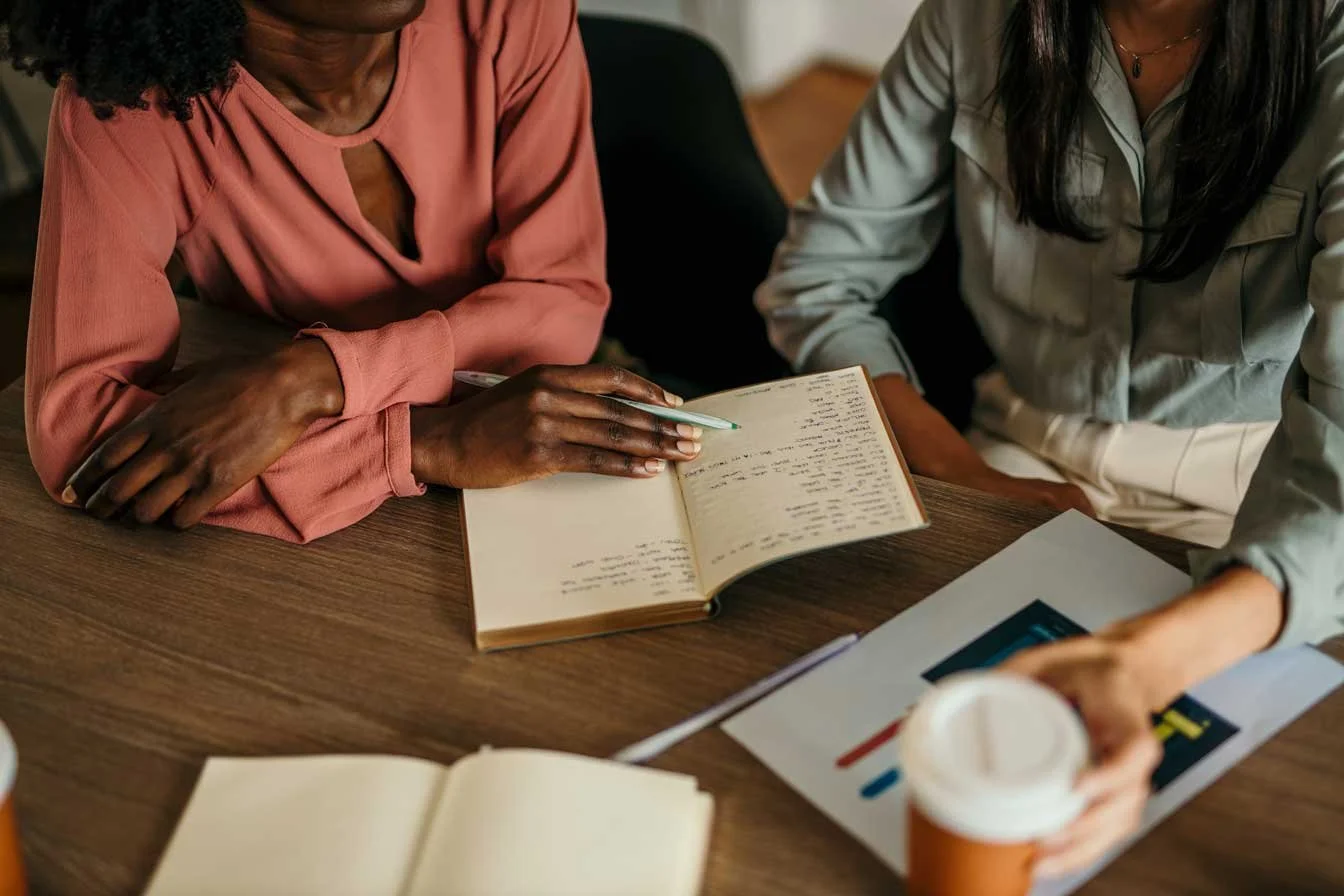 Two smartly dressed woman working together with an open notebook and coffee mug