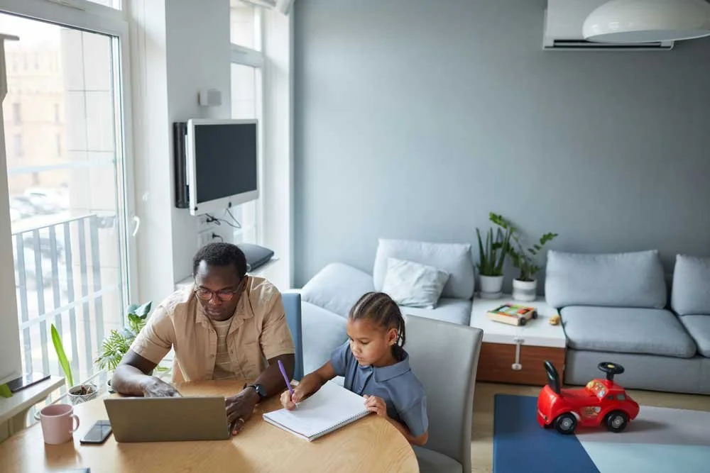 Black father working at his laptop and his son drawing at the desk in their living room.