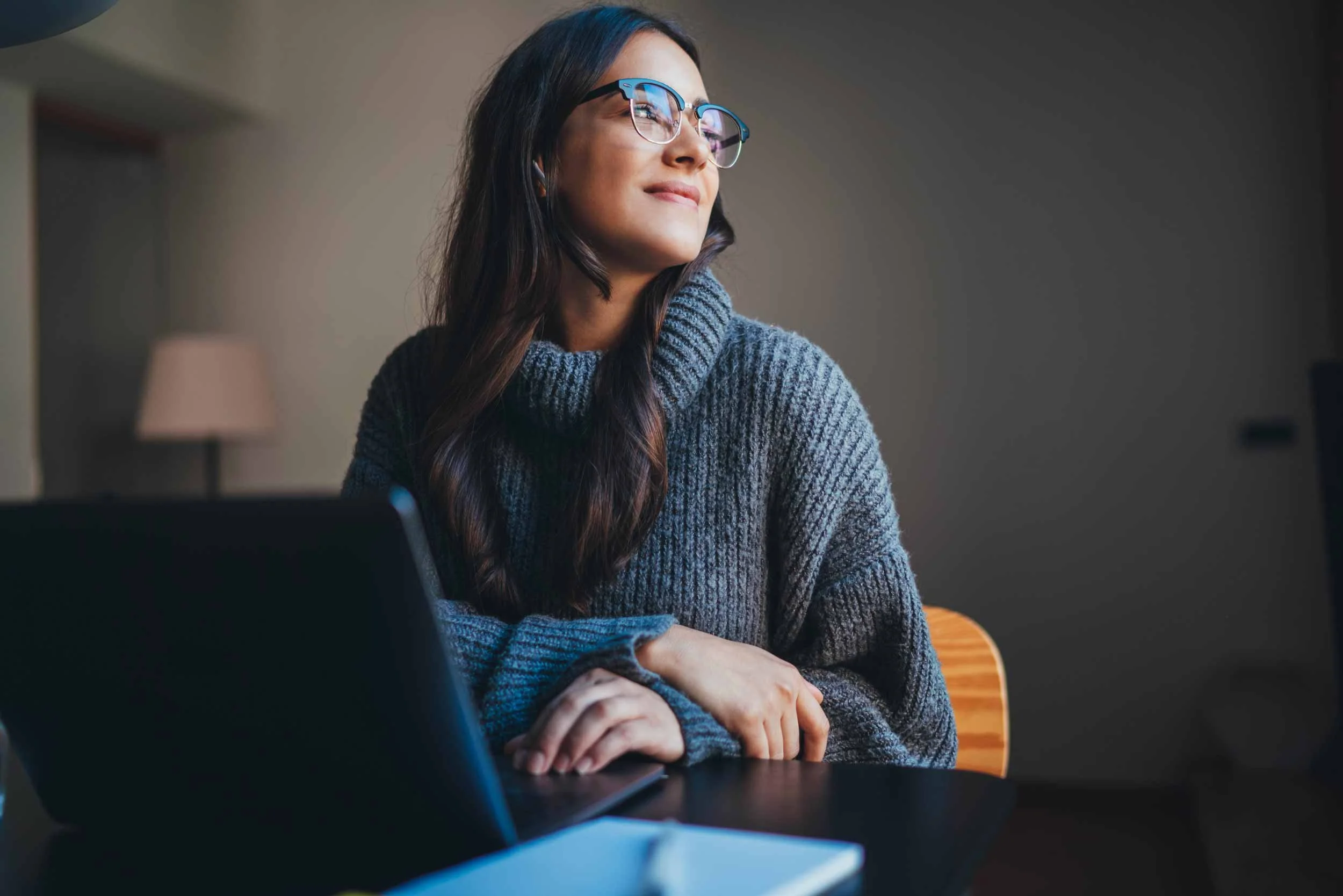 A VA in a gray sweater and glasses sitting at a desk with a laptop and notebook, looking upward with a relaxed expression in a softly lit indoor workspace