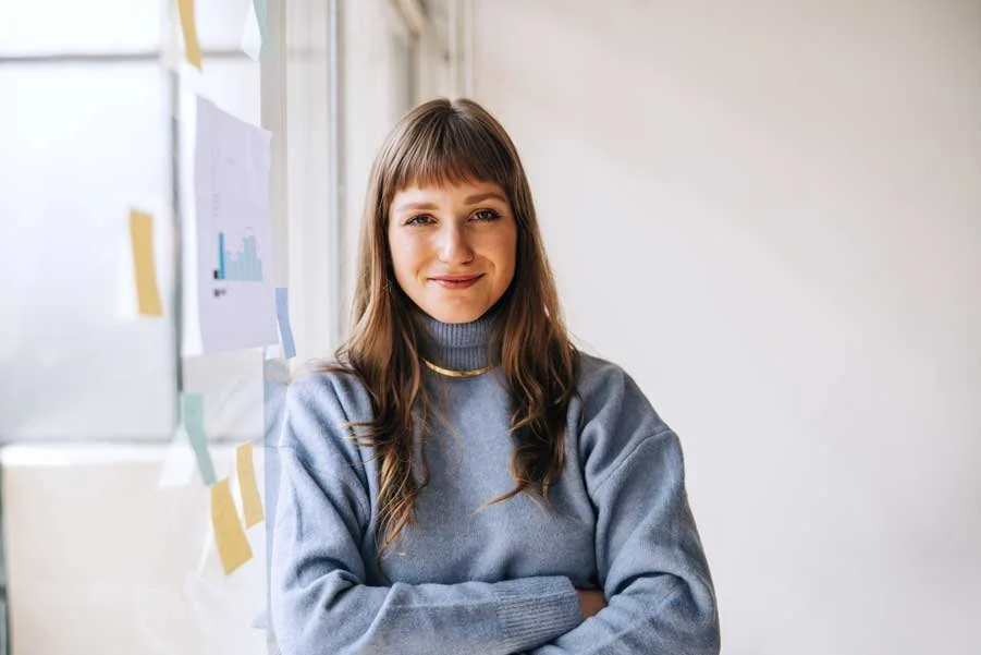 Blunt fringed woman stood smiling with folded arms next to a window covered in planning print-outs and sticky notes