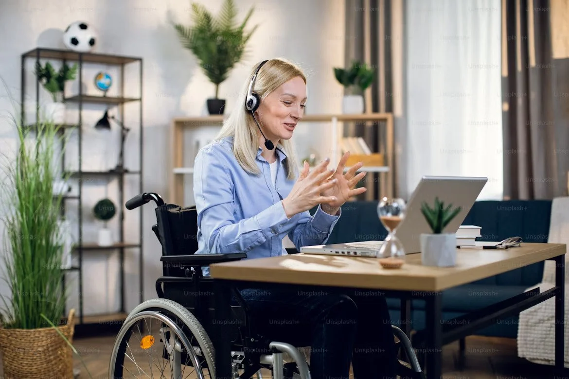 A VA in a wheelchair actively participating in a video call from a cozy home office, wearing a headset and gesturing while seated at a desk with plants, books, and a sand timer