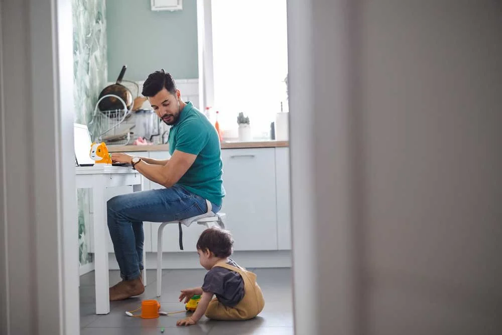 Barefooted father trying to work from home but distracted by his toddler playing next to him on the floor