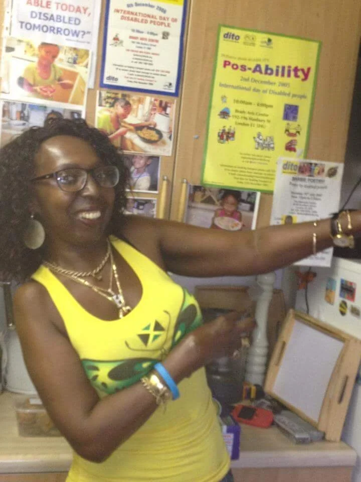 Smiling black woman donning a yellow Jamaican vest stood before a bright poster about endless possabilities for all
