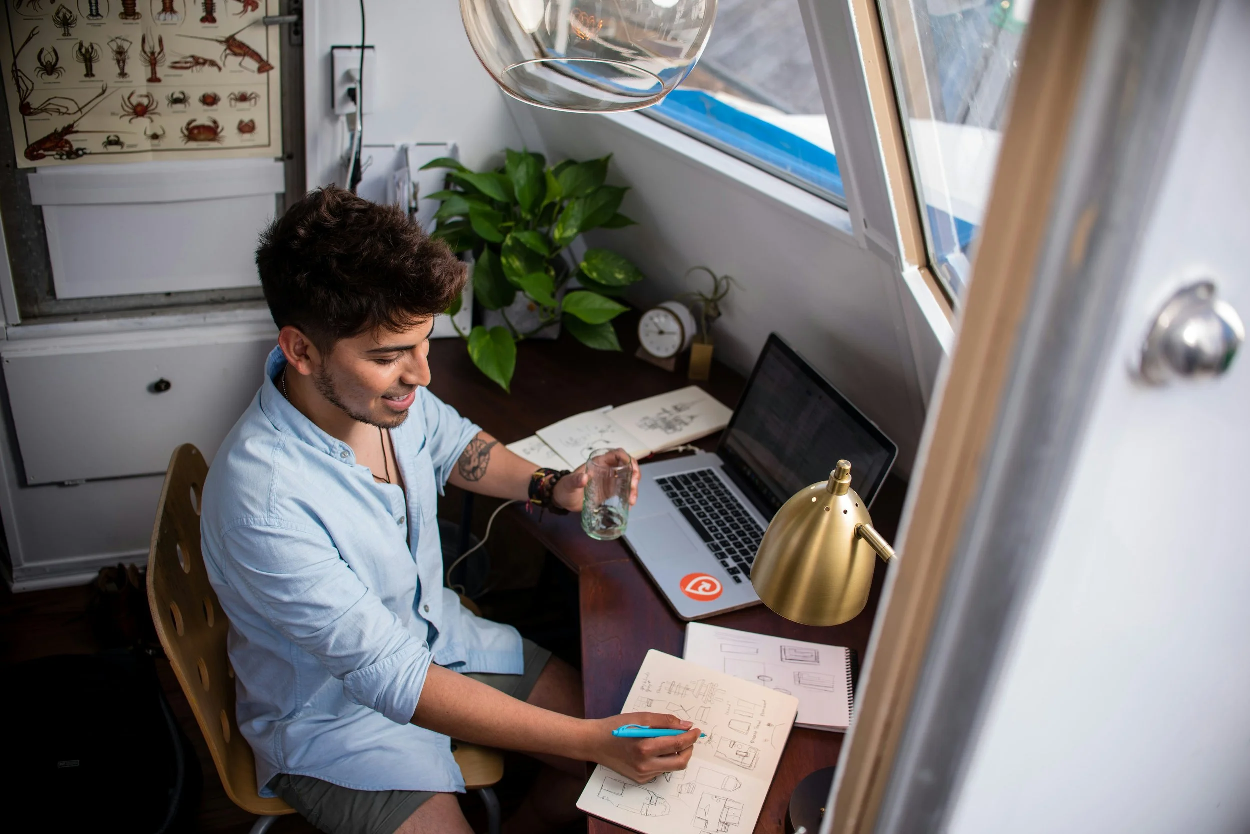 A web designer sketching wireframes in a notebook at a clean desk with a laptop, gold lamp, and marine-themed decor, suggesting a focused and creative design session in a nautical-inspired workspace