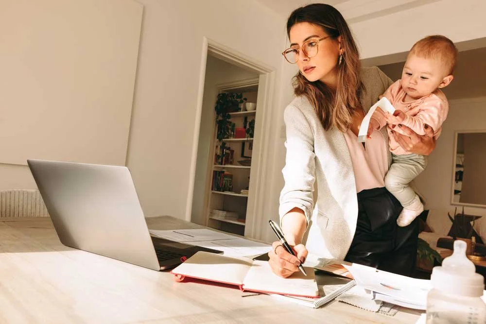 Stressed working mum typing on laptop standing with her baby on her hip