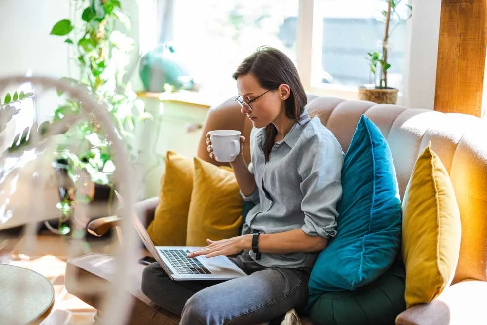 A VA working remotely on a laptop while seated on a colorful couch, holding a mug in a bright, plant-filled room with a relaxed yet focused atmosphere