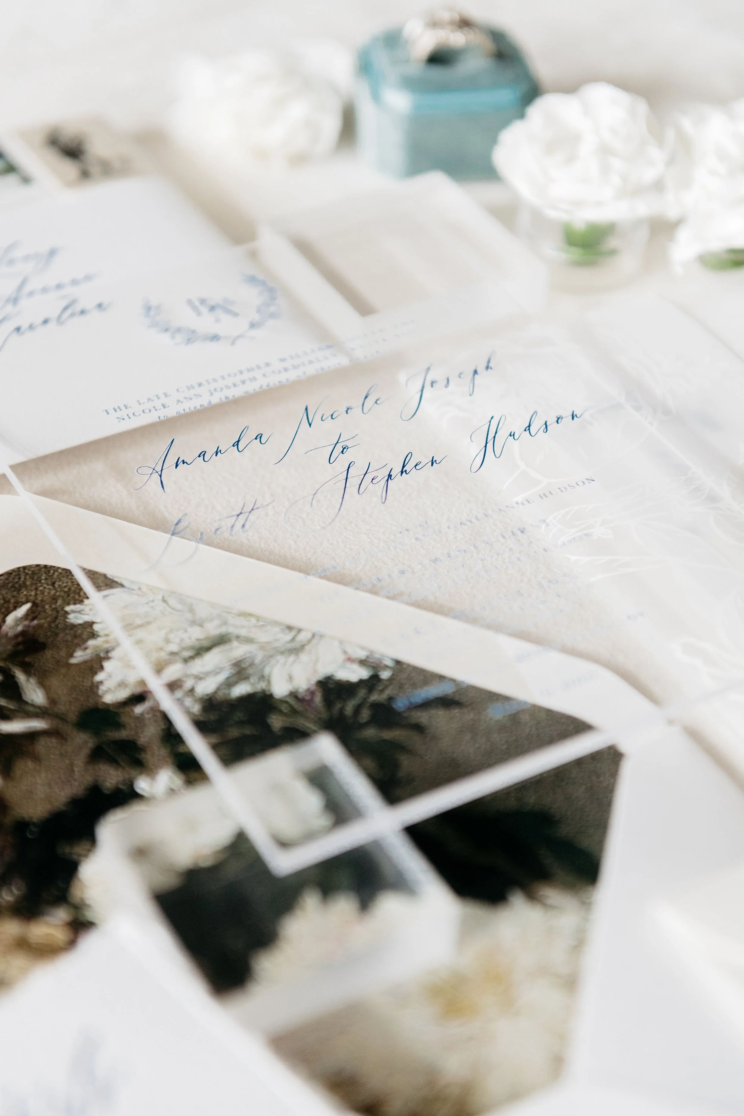 Wedding invitation set with floral details and calligraphy, accompanied by a ring box, decorative paper, and white flowers.
