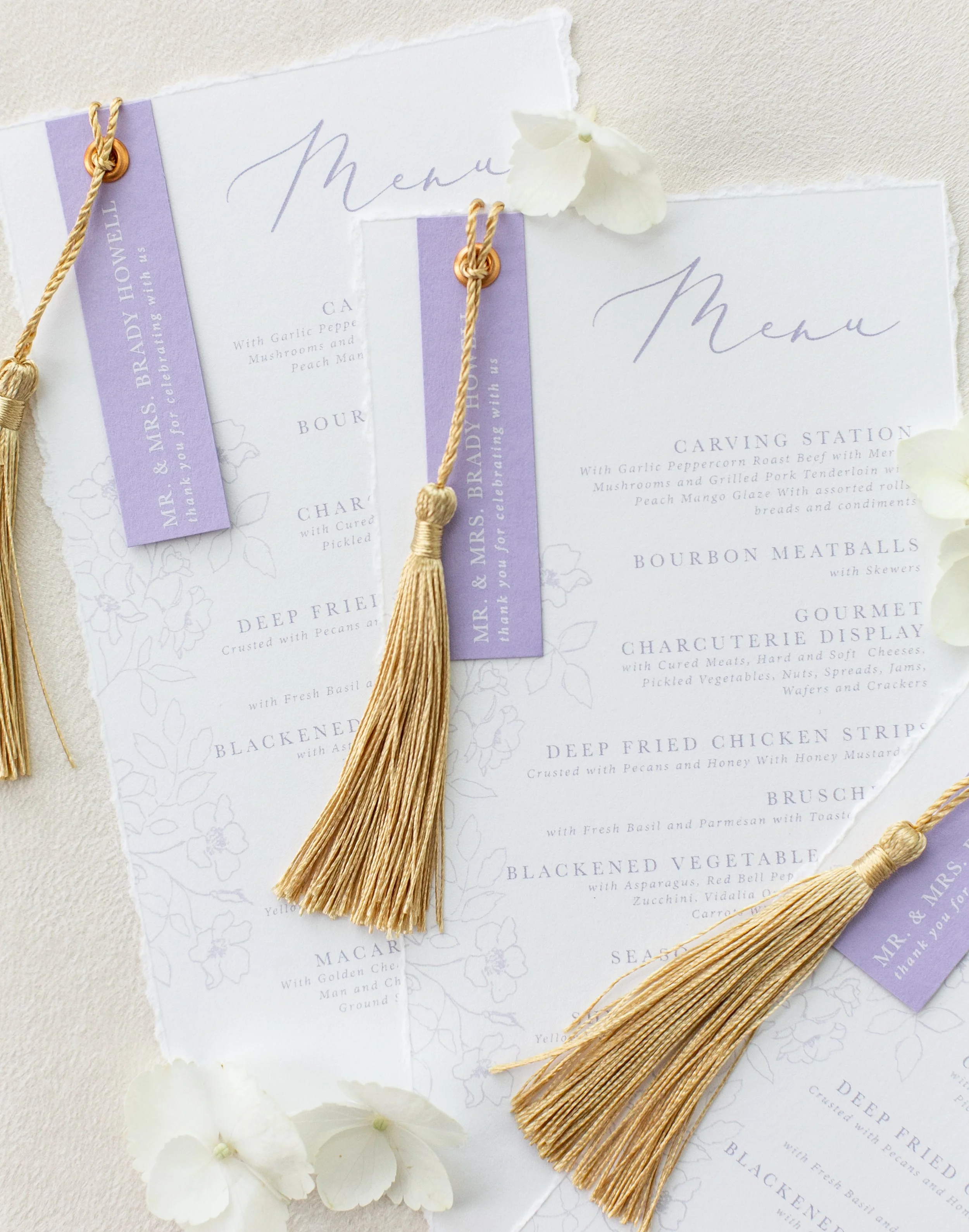 Elegant wedding menu cards with gold tassels and purple tabs, featuring floral designs, resting on a light textured surface, adorned with white flower petals.