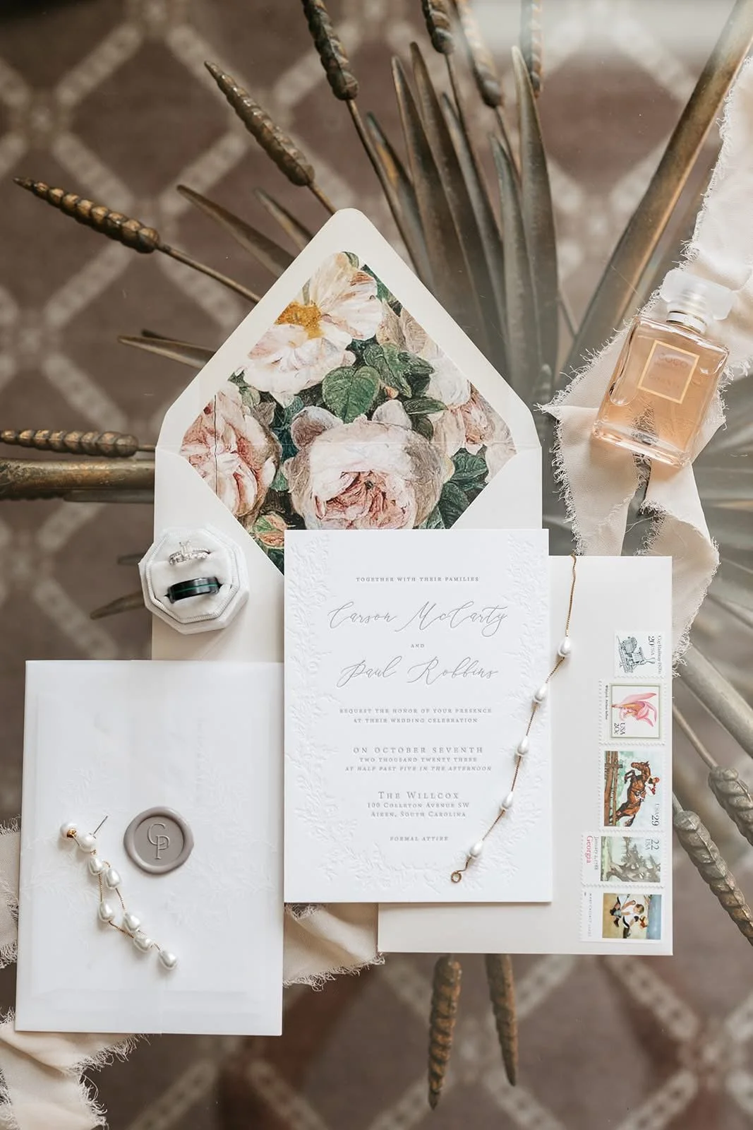 Elegant wedding invitation set with floral envelope lining, pearl bracelet, ring in a jewelry box, vintage stamps, and a bottle of Chanel perfume on a textured surface.