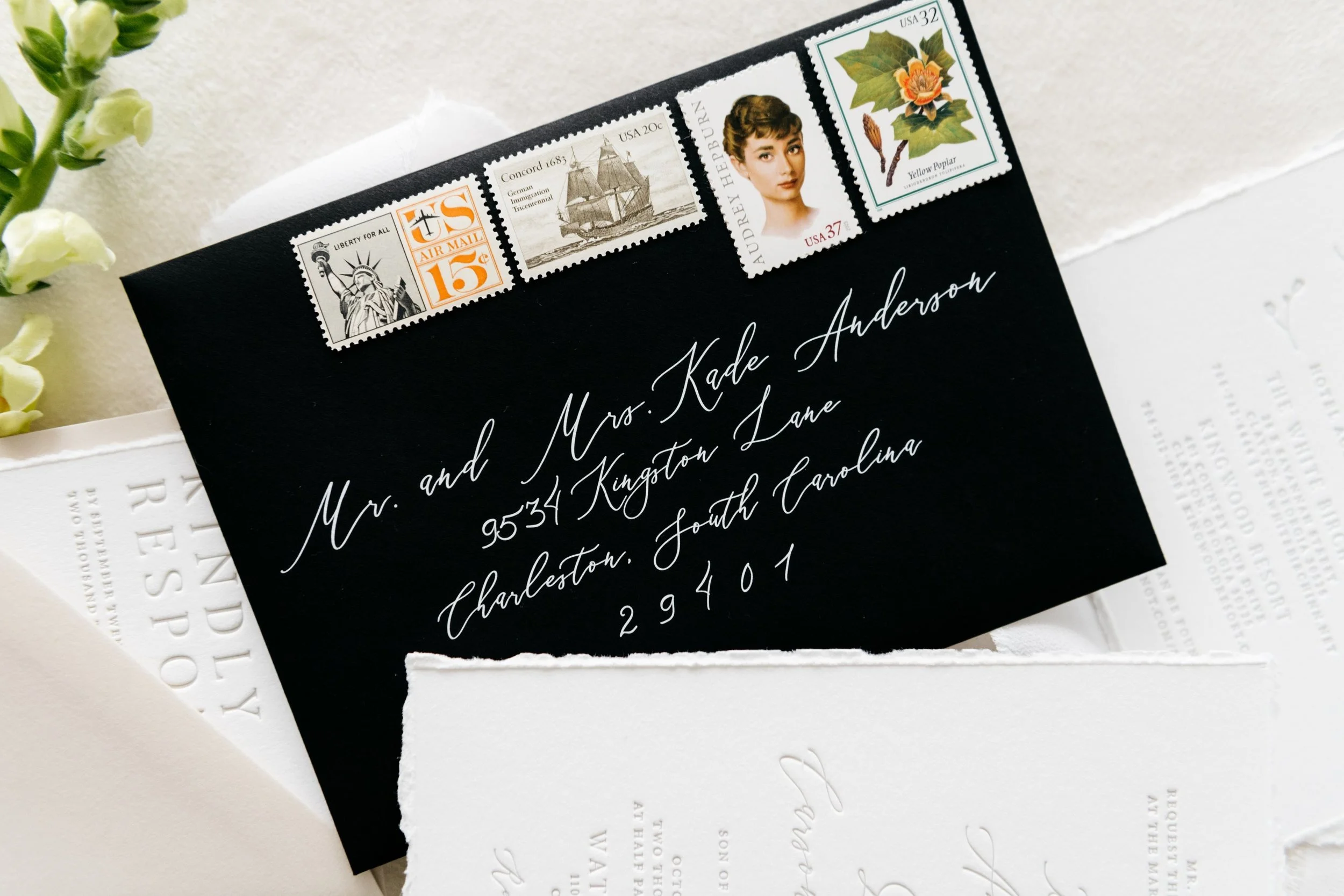 Black envelope with elegant white script handwriting, Charleston, South Carolina, adorned with various vintage postage stamps, including ones with the Statue of Liberty and flowers.