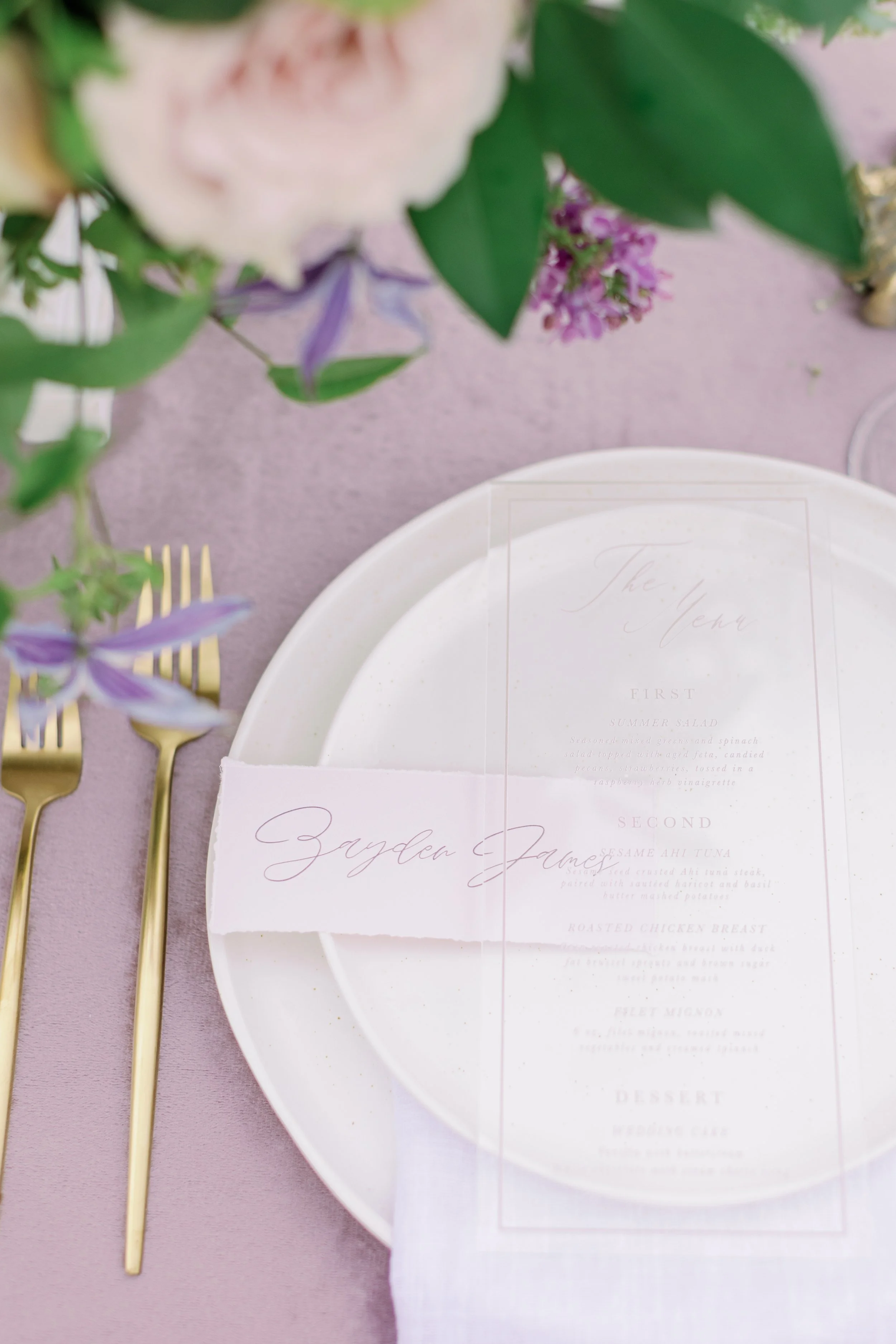 Wedding table setting with a white plate, a menu, and a place card with elegant script on a purple tablecloth, adorned with flowers and gold cutlery.