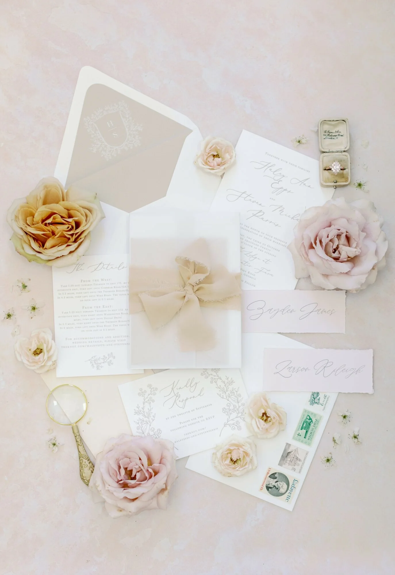 Flat lay of elegant wedding invitations surrounded by blush roses, a magnifying glass, vintage stamps, and a ring in a box. Phoenix, Arizona