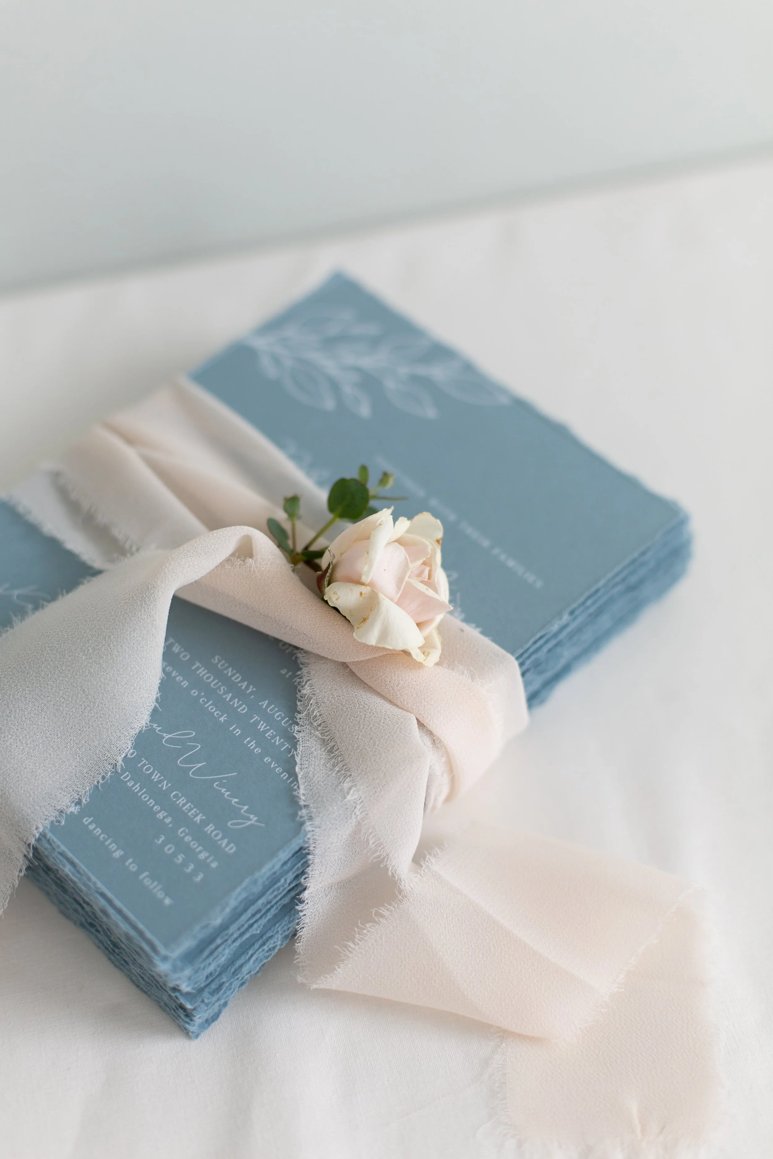 Stack of dusty blue wedding invitations tied with sheer ribbon and decorated with a small white rose. Charleston, South Carolina