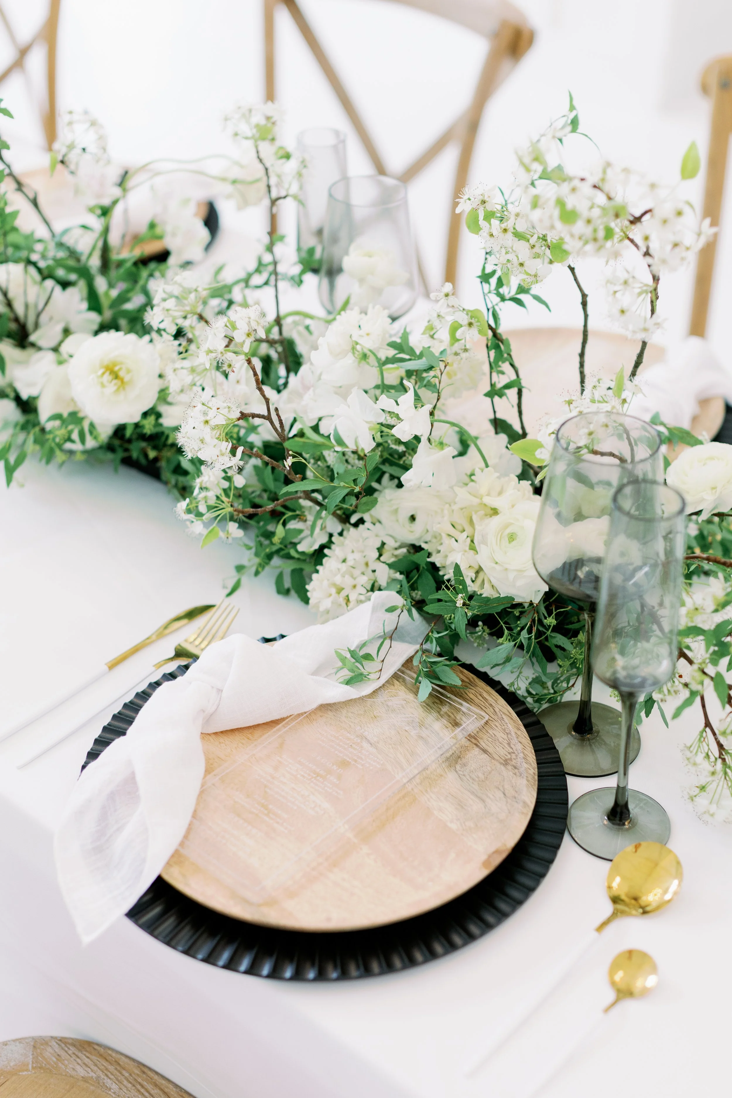 Elegant table setting with a wooden plate on a black charger, white napkin, gold utensils, and dark glasses. The centerpiece features white flowers and greenery.