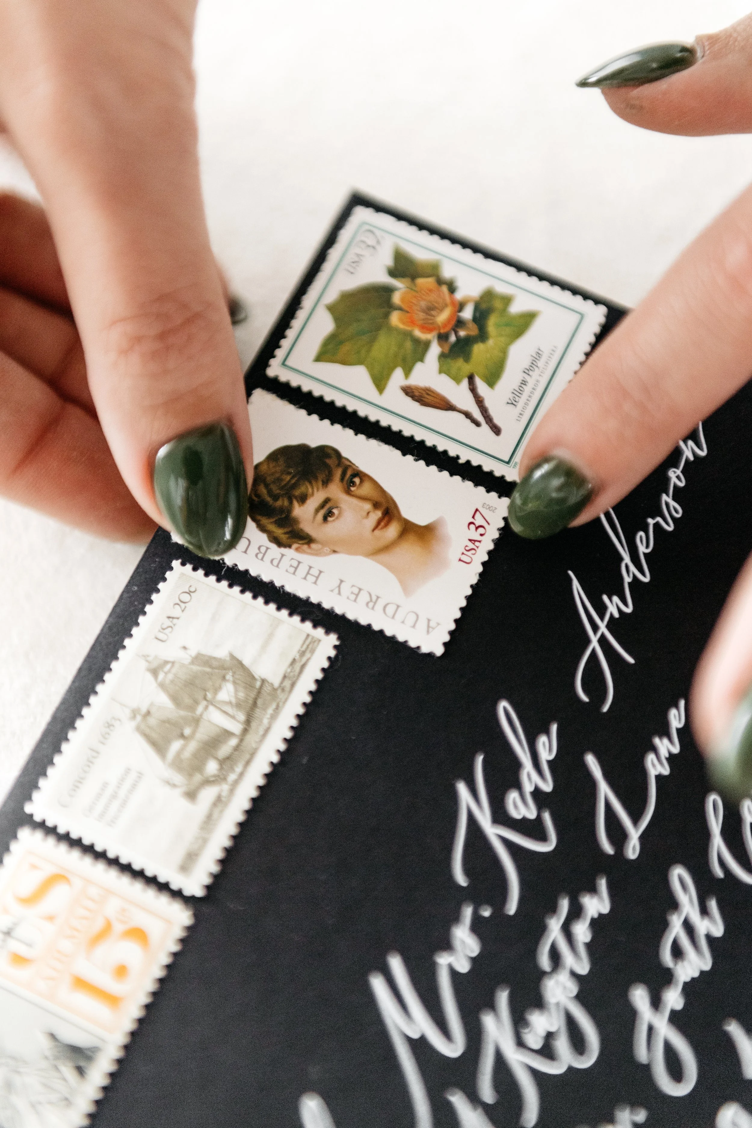 Hands placing stamps on a black envelope with calligraphic writing; stamps include an illustrated flower, a historical figure, and a ship.