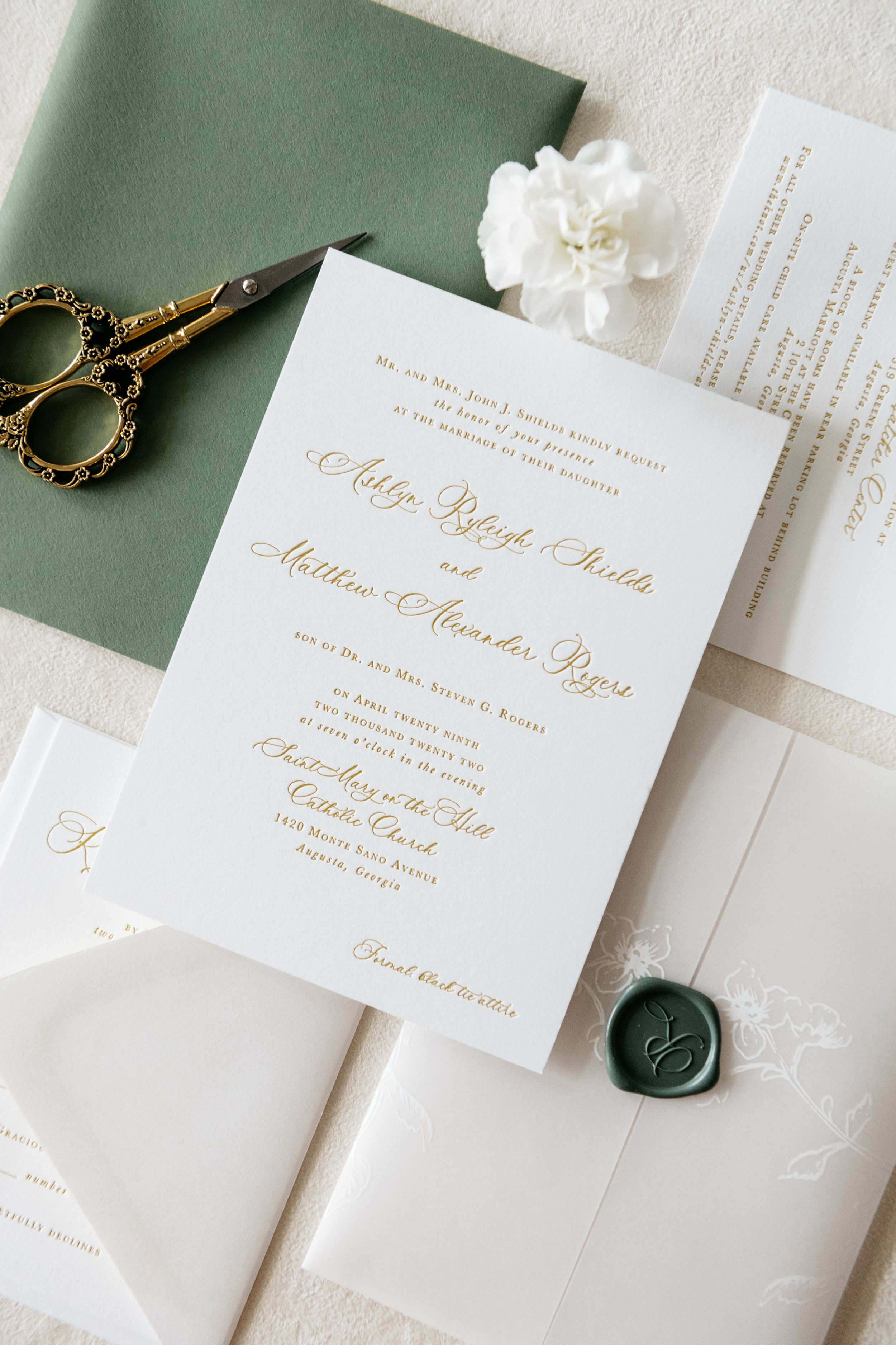 A set of elegant wedding invitations featuring gold script on white paper, accompanied by a green envelope, ornate gold scissors, a sealed envelope with a floral design and wax seal, and a white carnation flower. Charleston, South Carolina