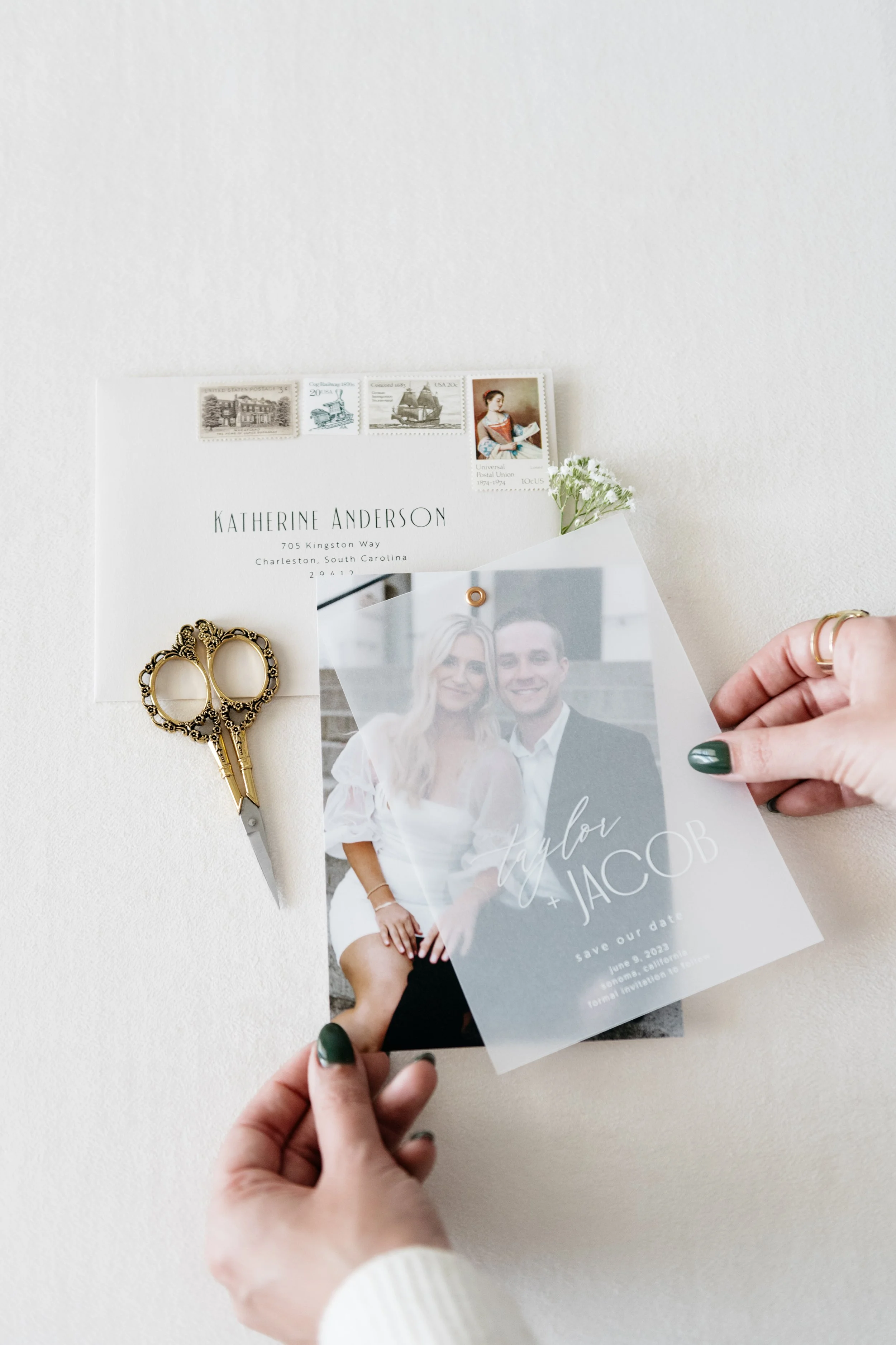 A hand holding a save-the-date card featuring a couple's photo, partially overlaid by a translucent sheet with the text "taylor + JACOB" and event details. Nearby is an envelope with stamps and a pair of ornate scissors on a white surface.