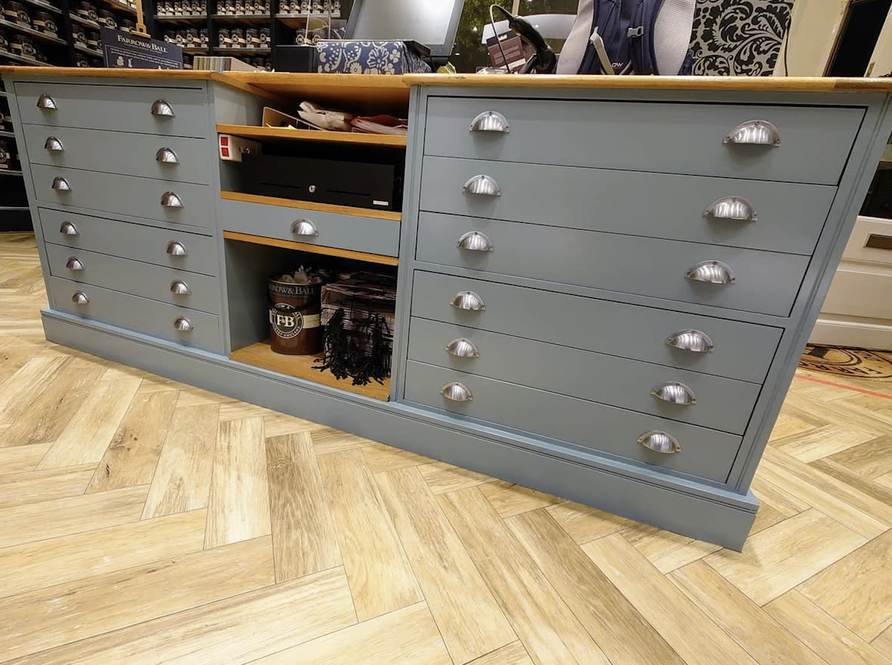 A light blue wooden dresser with silver half-moon drawer handles, featuring multiple drawers, open shelves with office supplies, and a wood top surface, positioned on a light wood herringbone floor.
