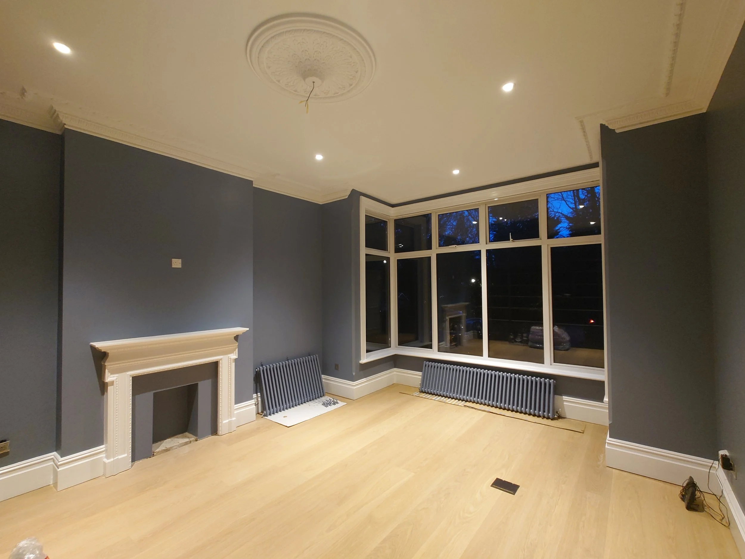 Interior of a room with blue walls (Farrow & Ball Selvedge Dead Flat), a white fireplace, large bay window, and wooden flooring.