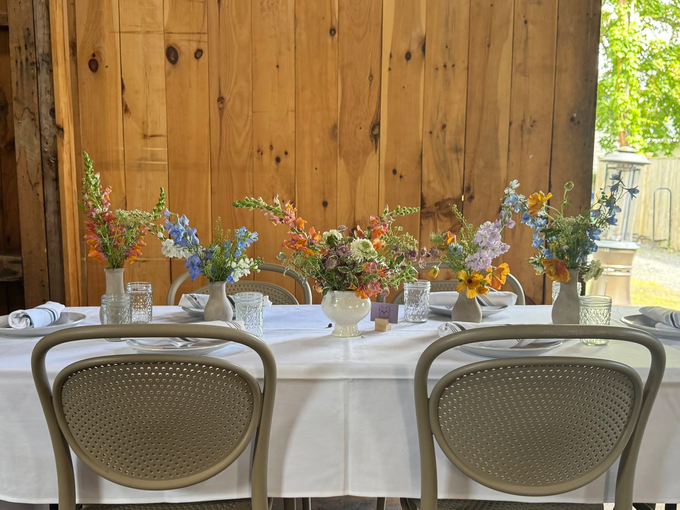 Dining table set with flower arrangements in vases and glassware, in a room with a wooden wall and windows with greenery outside.