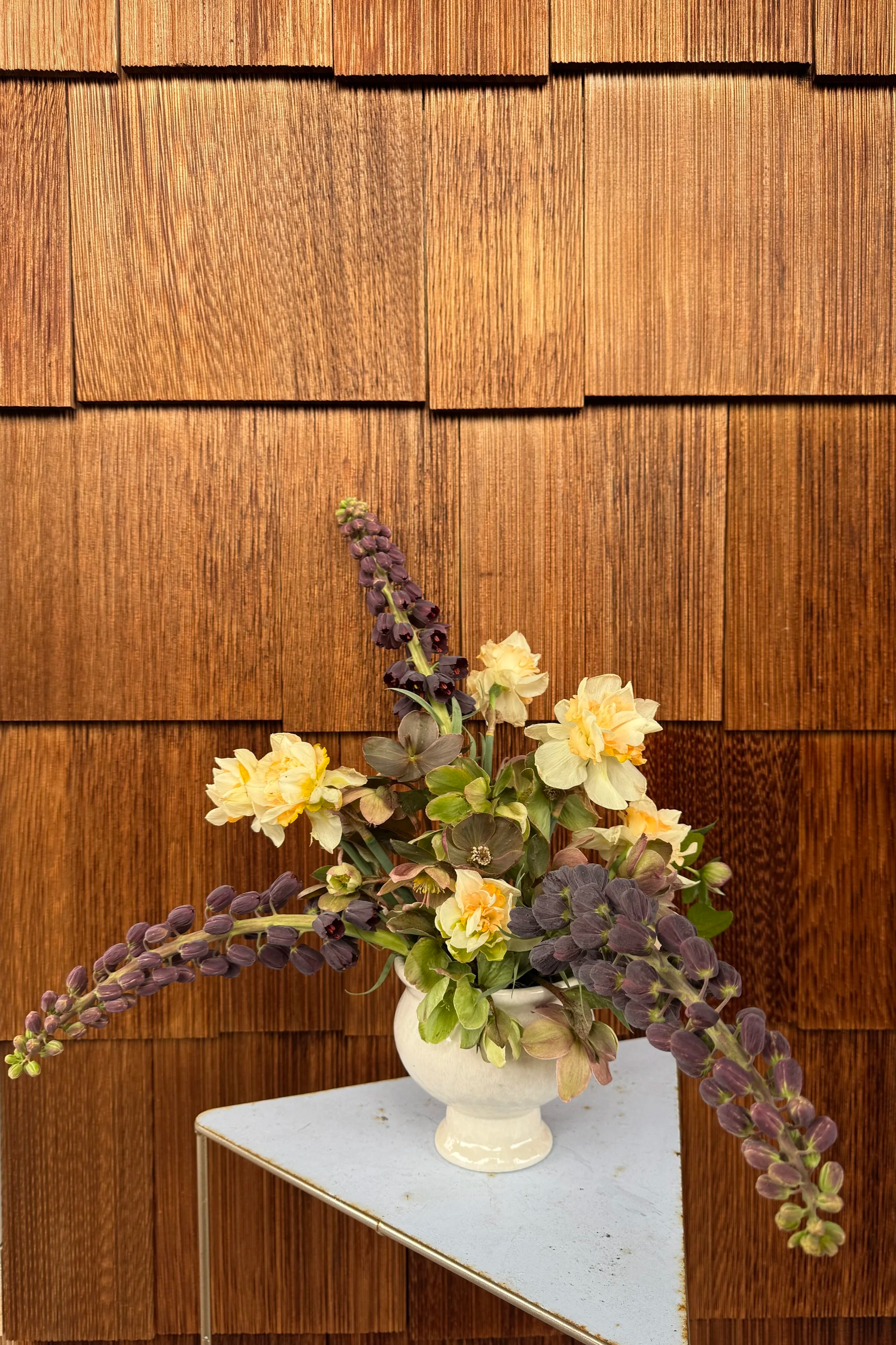 A floral arrangement with purple, cream, and green flowers in a white vase on a small white table, set against a wooden shingle wall.