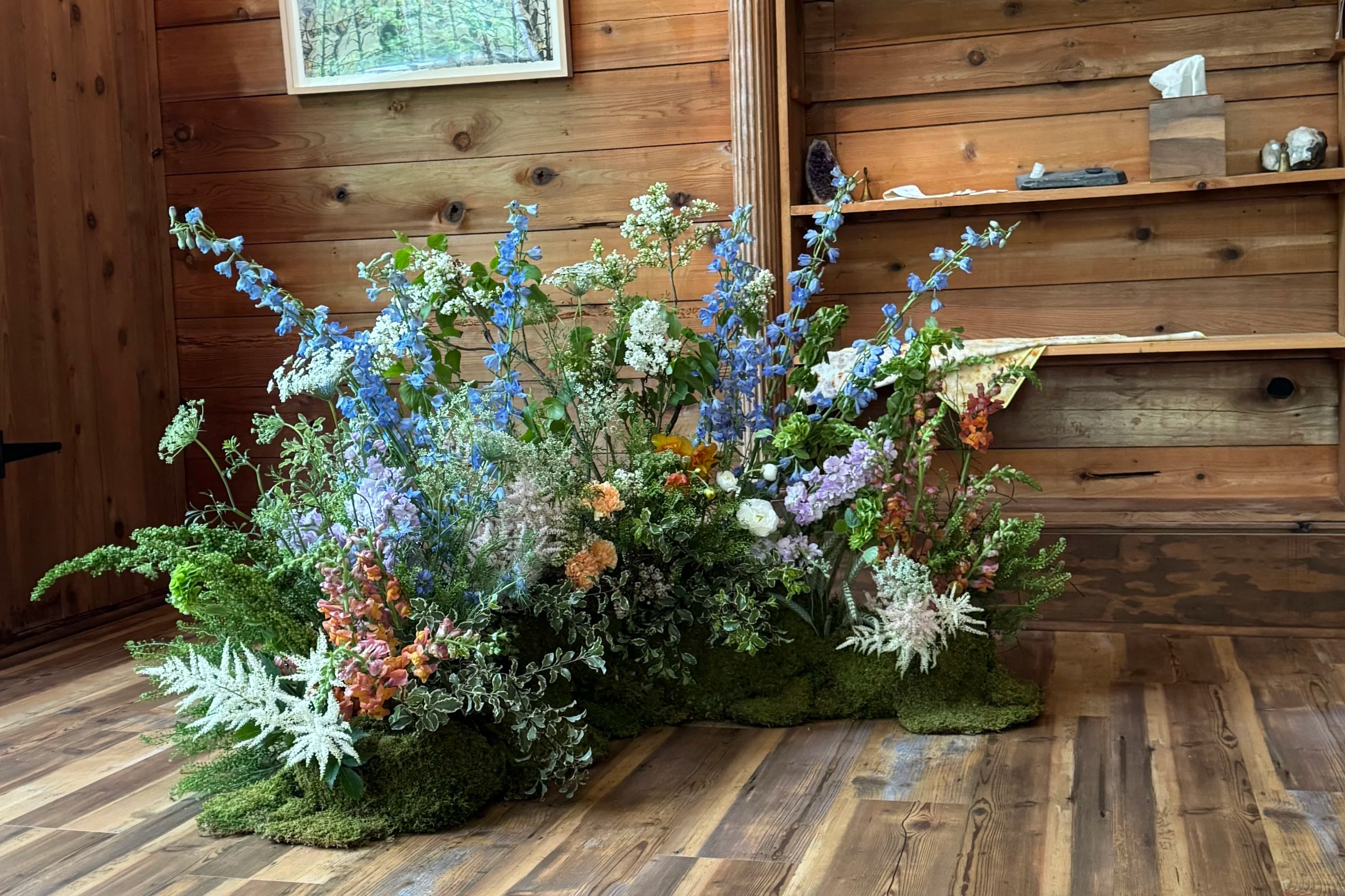 A large arrangement of colorful flowers is placed on a wooden floor inside a room with wood-paneled walls. The room features a framed artwork on the wall and some decorative items on built-in shelves.