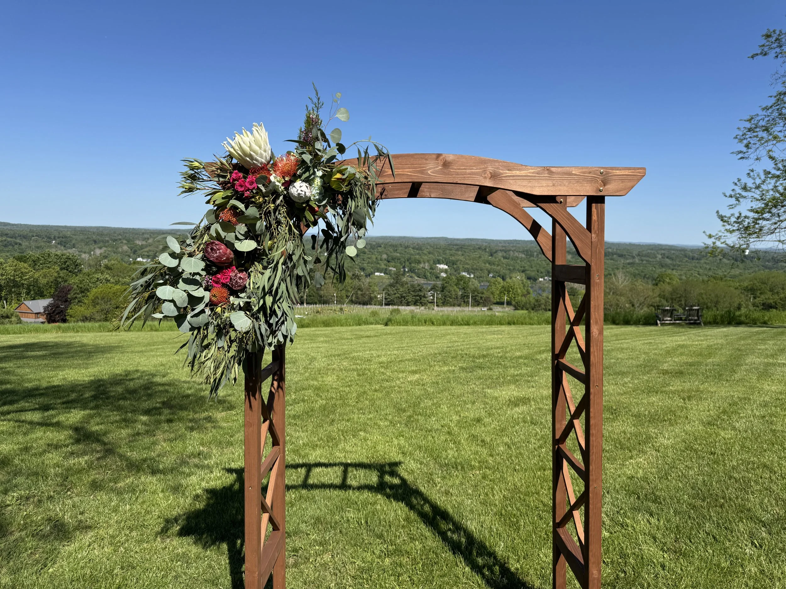 A wooden outdoor arch decorated with a floral arrangement including large white protea, pink roses, and greenery, set against a grassy field and a blue sky.