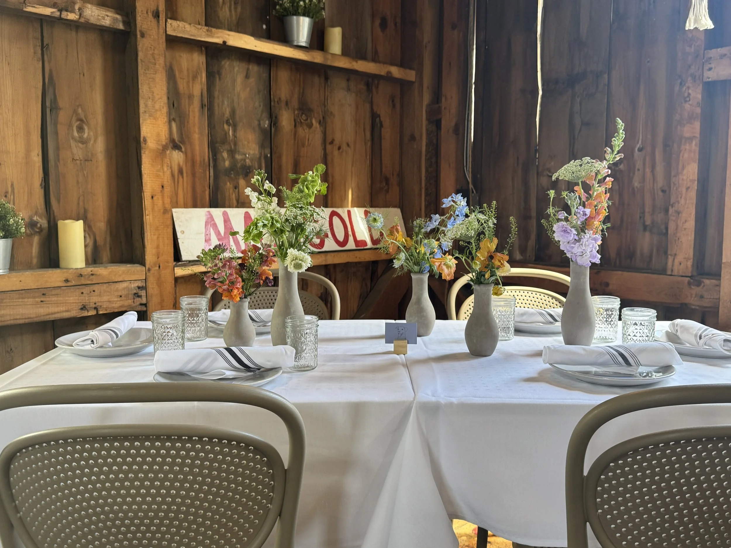 A rustic wooden interior decorated with potted plants and a vintage sign, featuring a table set with white tablecloths, vases of colorful flowers, glassware, and rolled napkins.