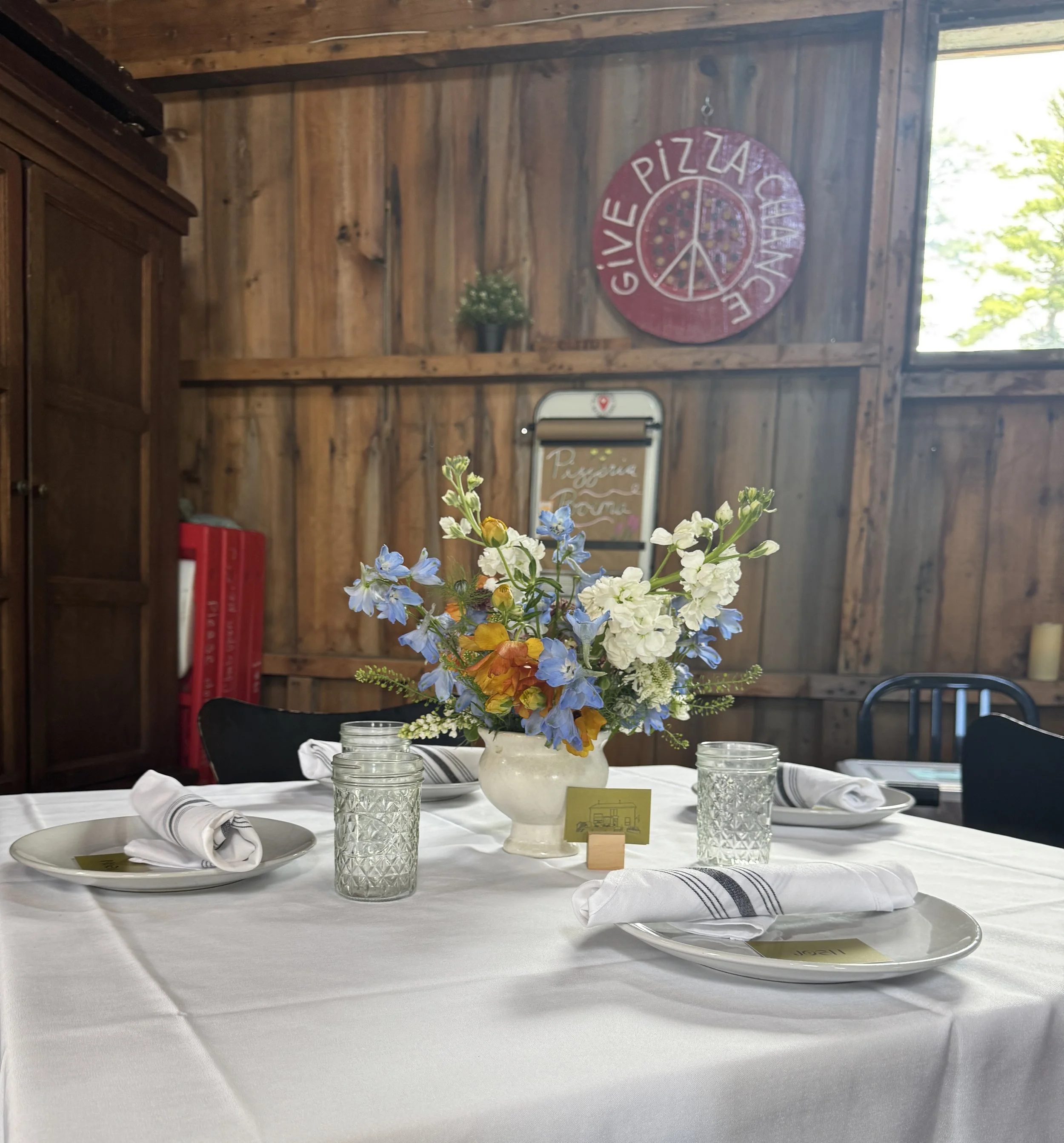 A dining table set for a meal with a floral centerpiece in a white vase, surrounded by plates, wine glasses, and cloth napkins, with a wood-paneled wall and a large window in the background.