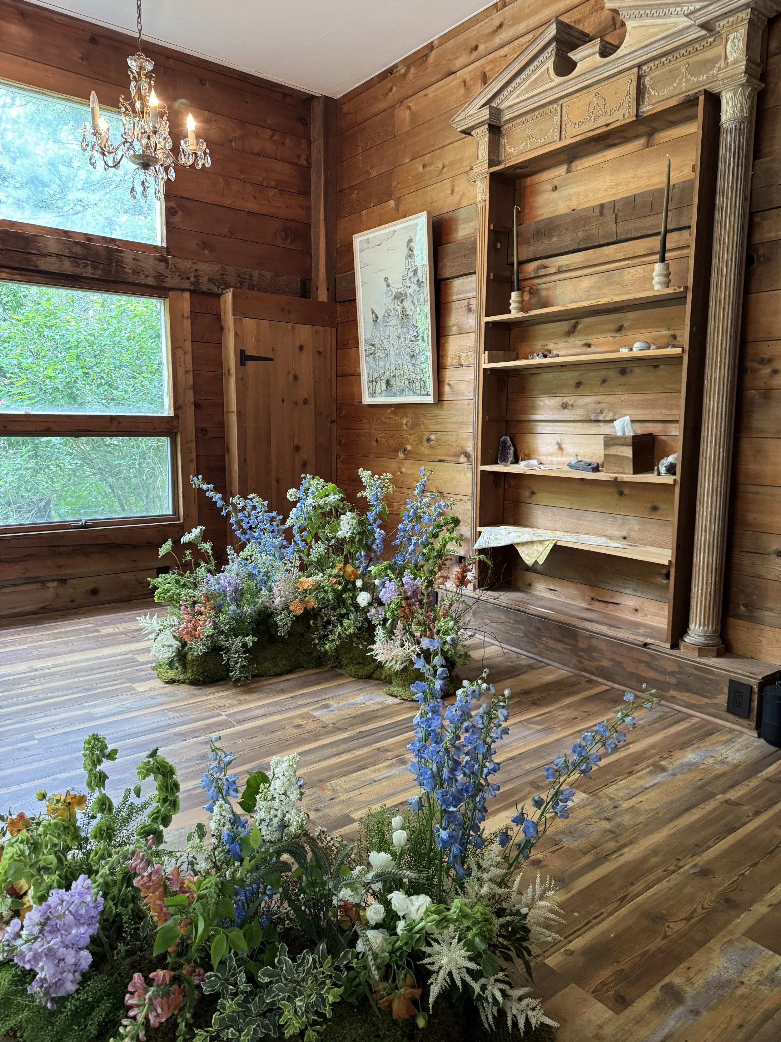 An interior wooden room with a large floral arrangement on the floor, featuring various flowers and greenery. There is a wooden bookcase on the wall, a framed artwork, wood-paneled walls, and natural light coming from a window.