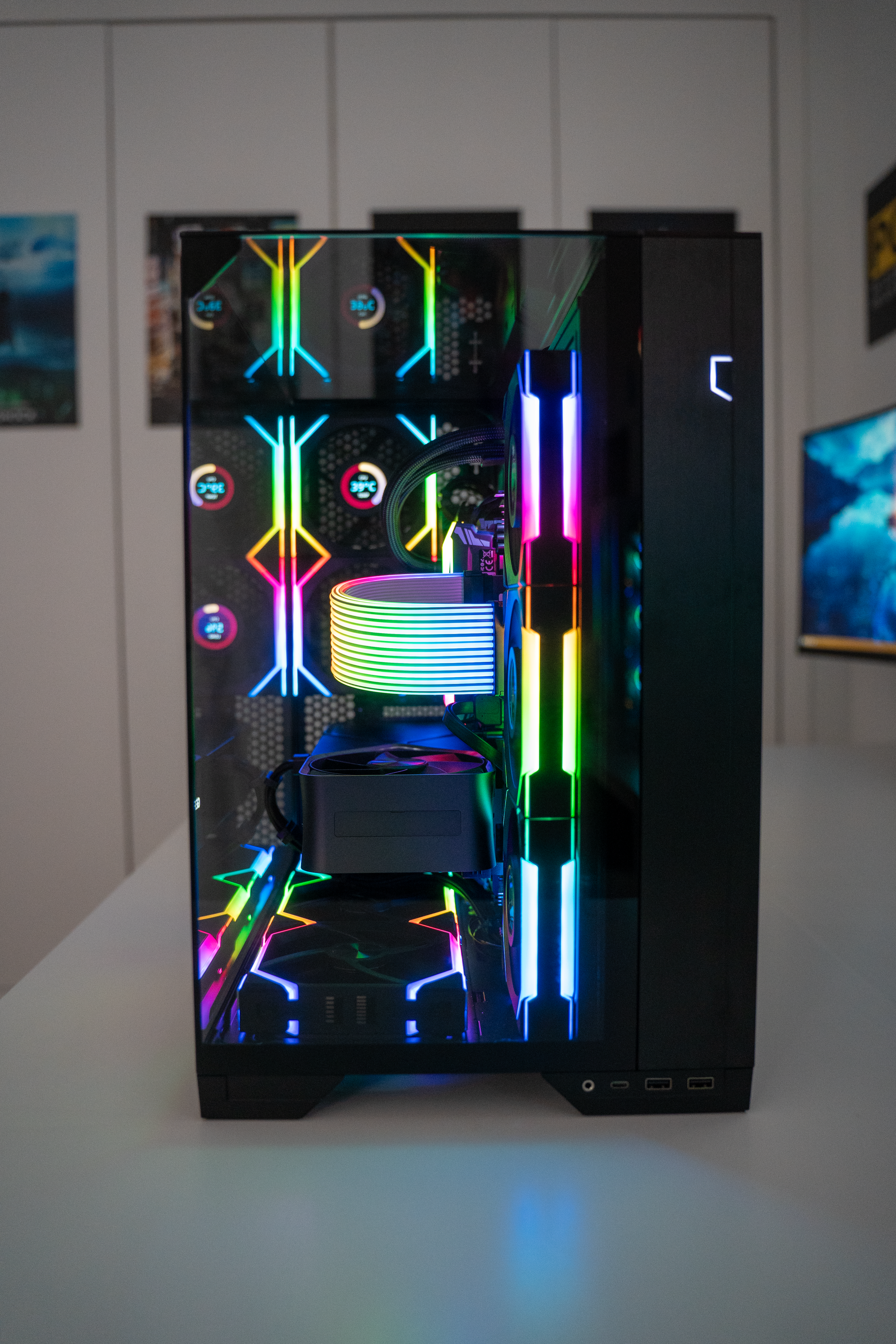 A gaming computer with colorful RGB lighting inside a glass case, positioned on a white surface with a background of posters on the wall.