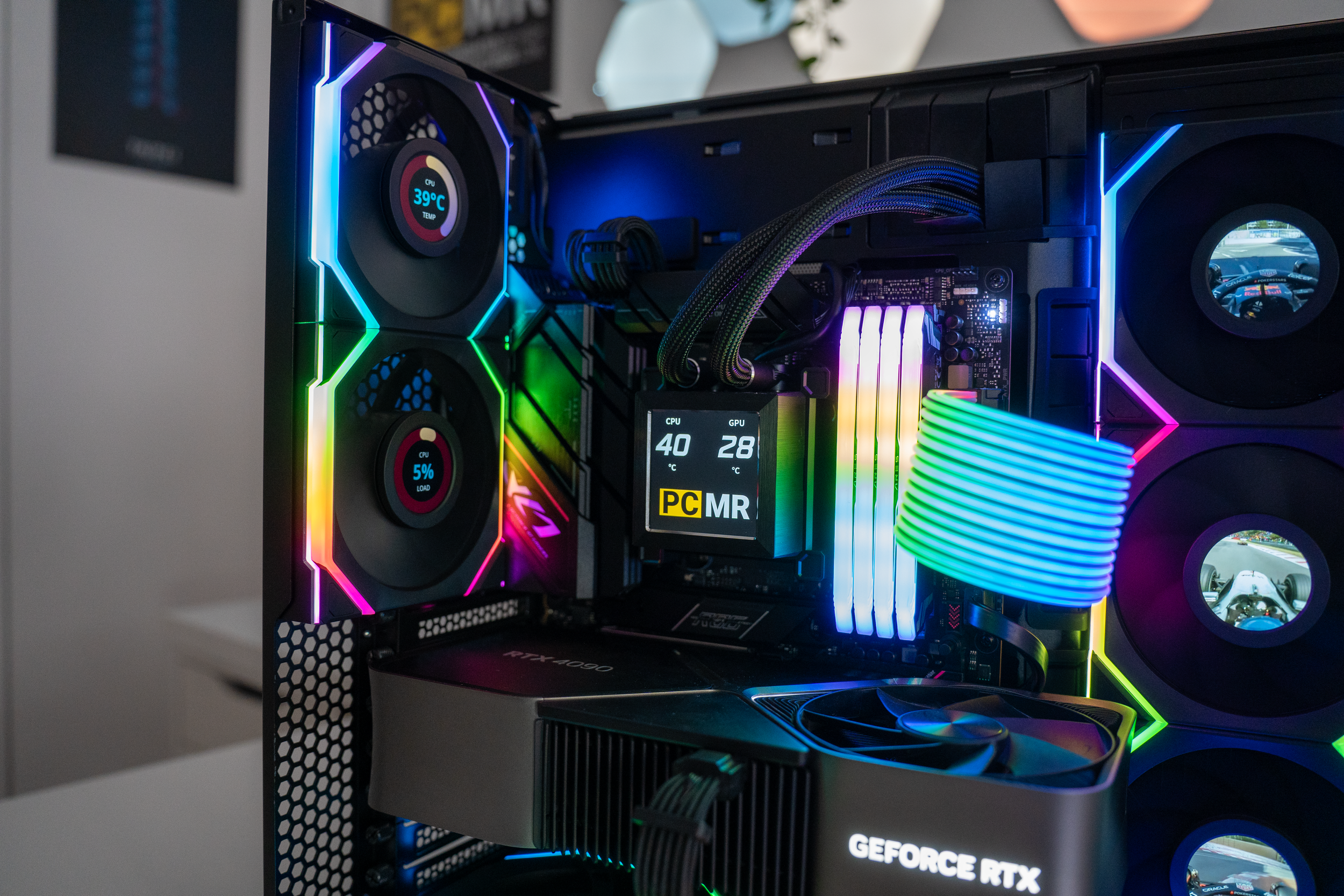 Inside a gaming PC with RGB lighting, featuring two large fans on the left, an RGB RAM module, a liquid cooling system, an NVIDIA GeForce RTX graphics card, and a small display showing CPU and GPU temperatures.