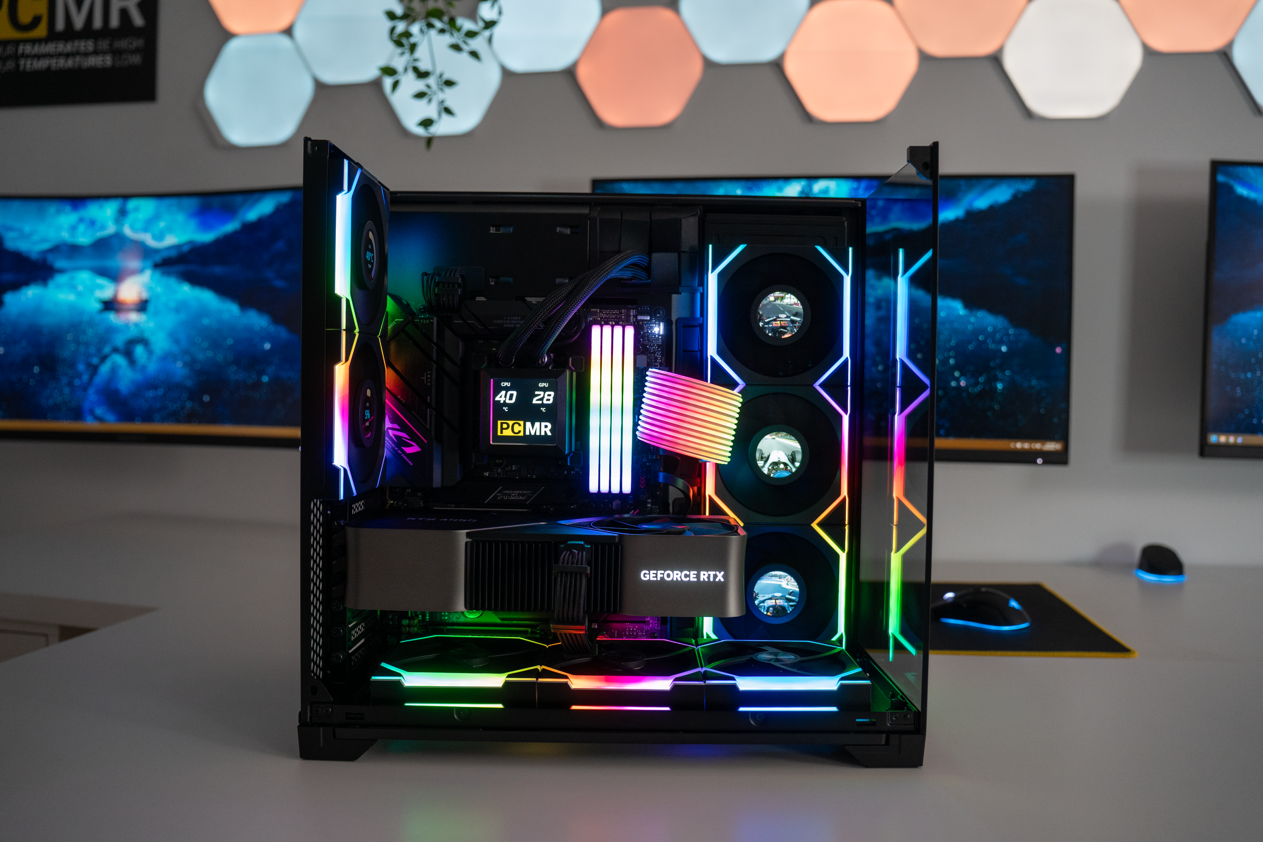 Open gaming computer case with RGB lighting showing CPU, GPU, fans, and memory modules, set on a white desk with dual monitors and a mouse in a modern workspace.