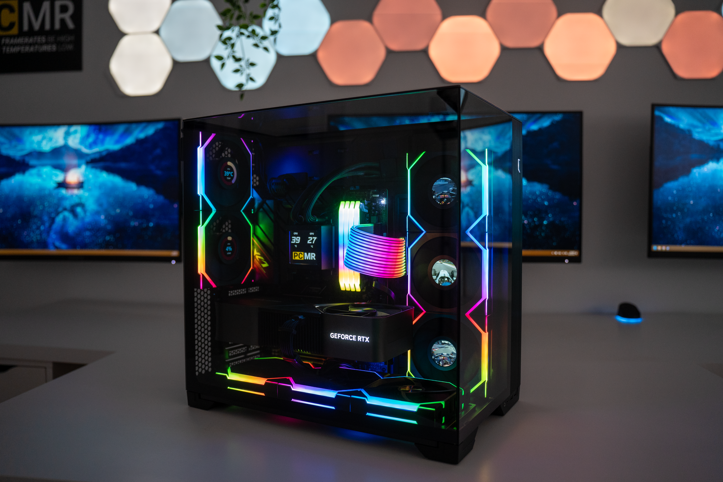 A gaming or high-performance computer with illuminated RGB lighting inside a transparent case, placed on a white desk with dual monitors displaying a dark, blue-toned landscape background in a modern, tech-themed room.