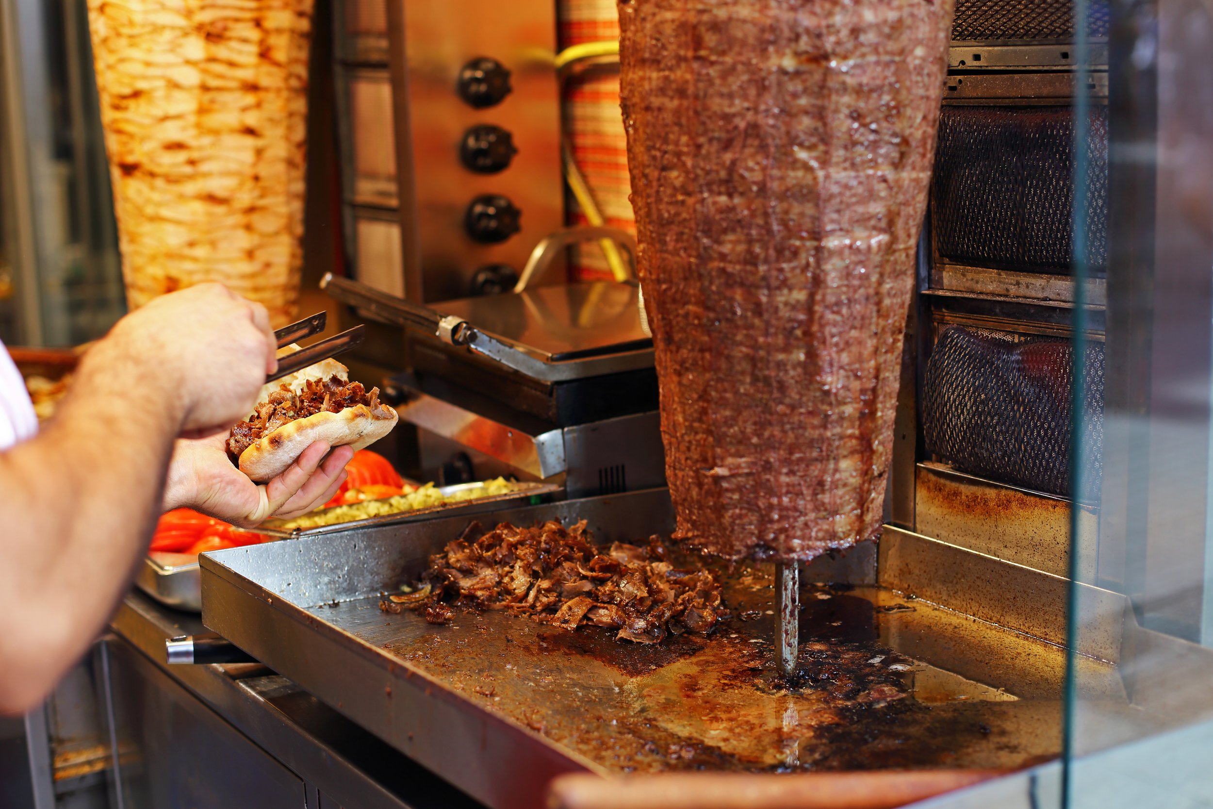 Person assembling a gyro sandwich with sliced meat on flatbread at a food cart, with a vertical spit of seasoned meat cooking behind them.