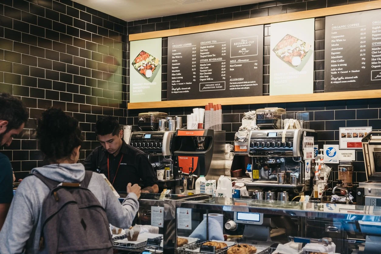 Customers ordering at a coffee shop counter with baristas preparing drinks, black tiled wall, coffee machines, and menu boards above.