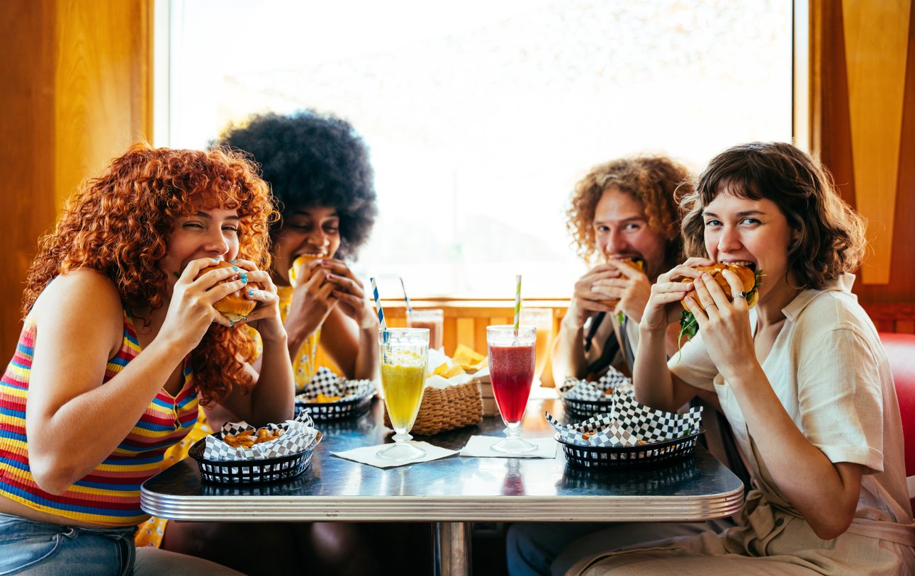 Four women sitting at a table in a restaurant, eating burgers and drinking colorful beverages.