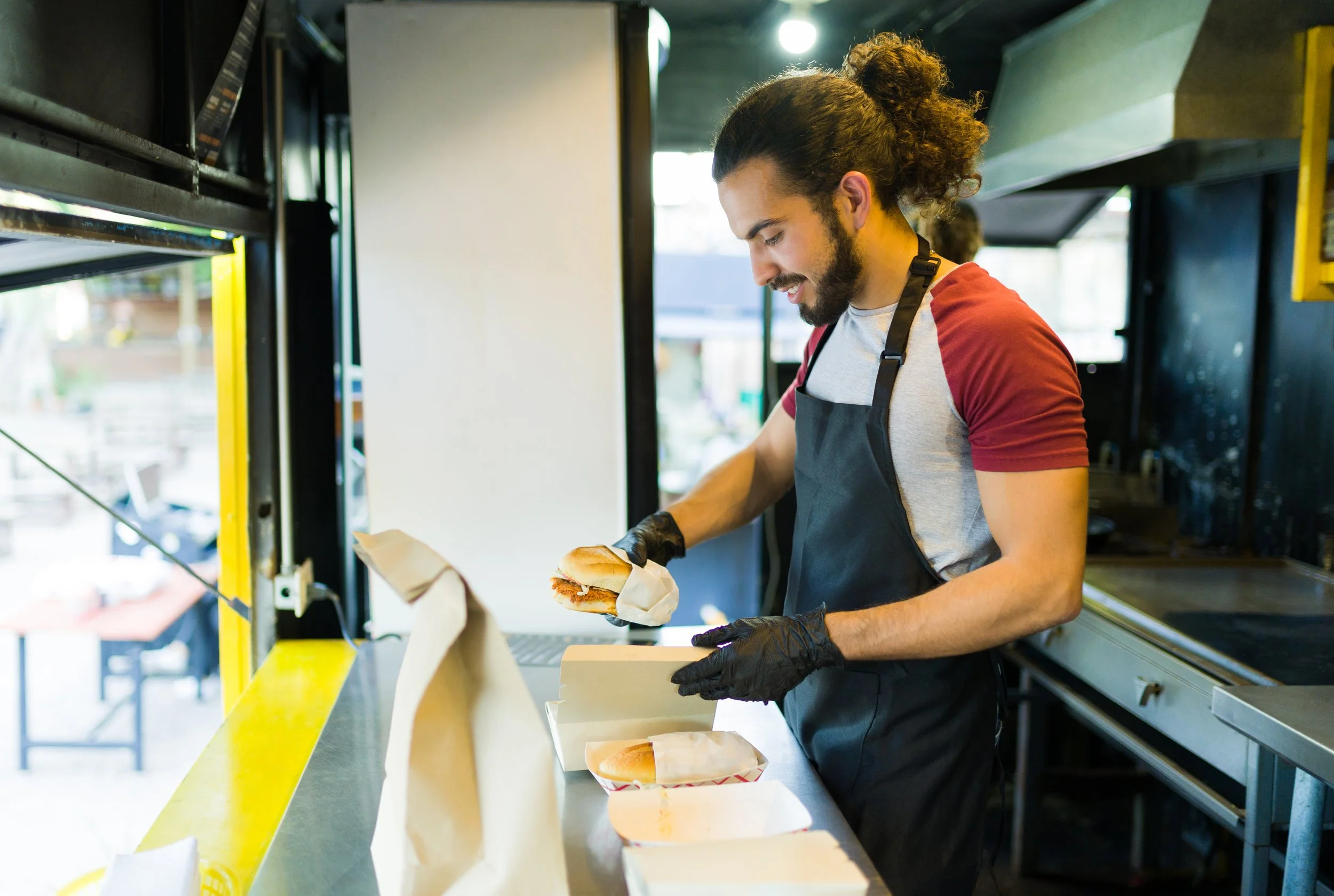 A young man with curly hair, wearing a gray T-shirt and black apron, prepares a sandwich in a food truck. He is holding a sandwich wrapped in paper and smiling.